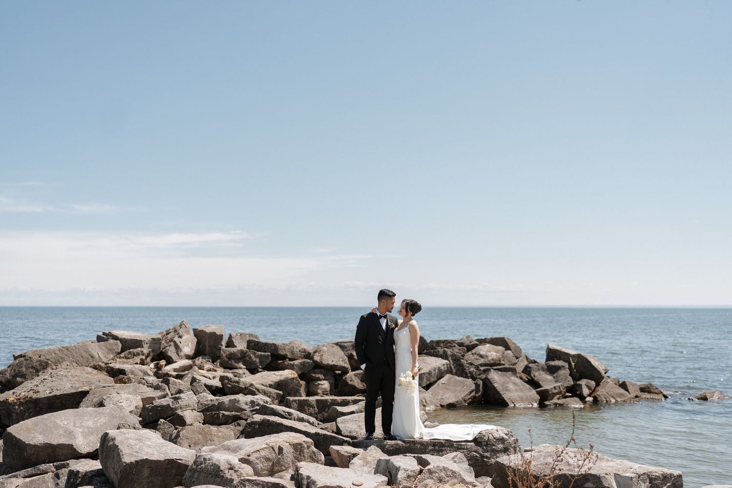 A couple stands together on a rocky ocean shore under blue skies on their wedding day.