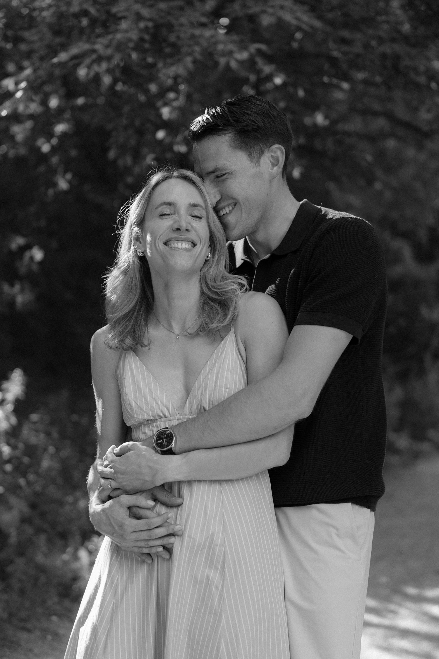 Black and white photo of a happy couple embracing outdoors, woman in sundress smiling, man in polo shirt hugging her.