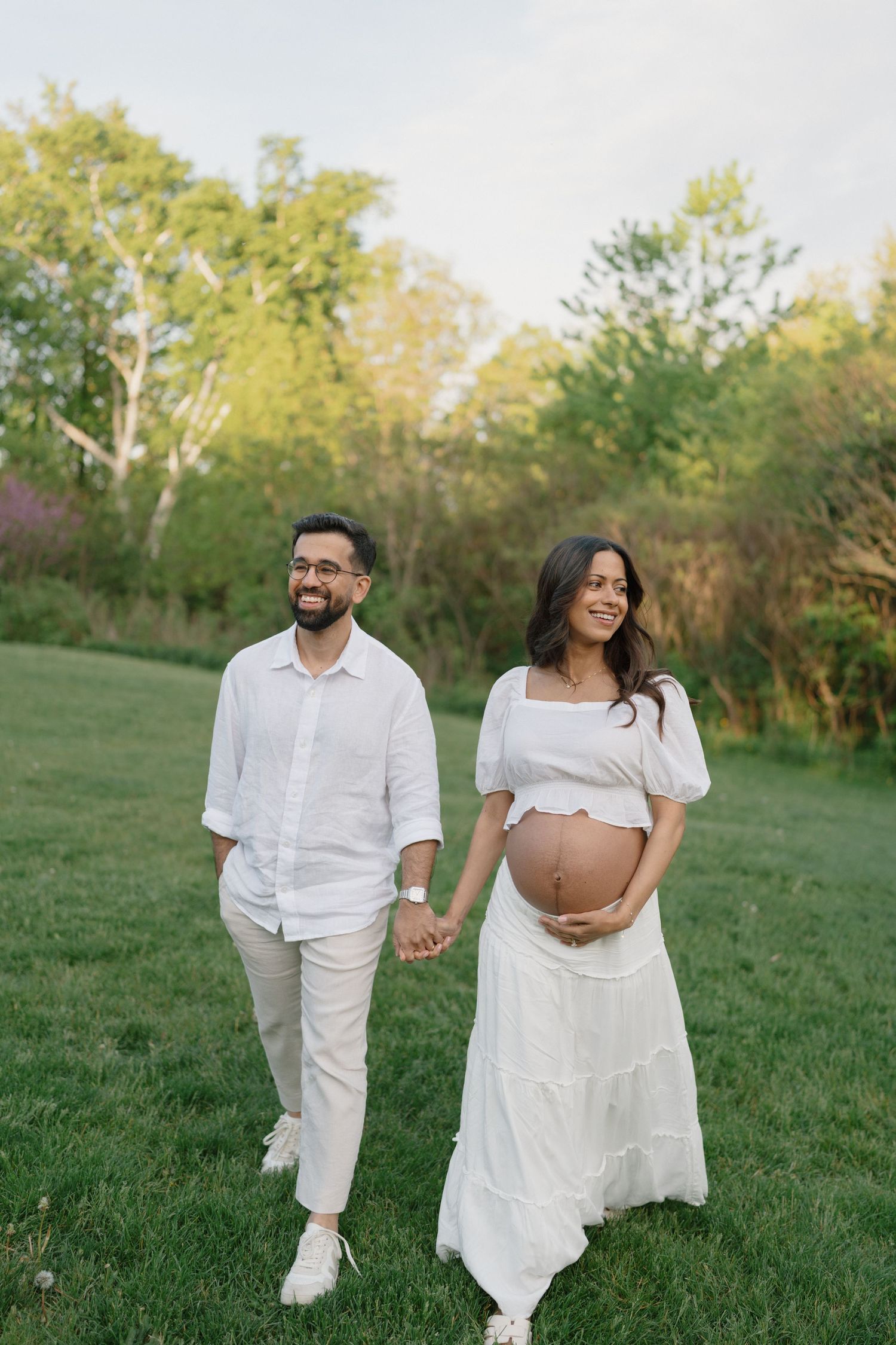 Happy expecting couple in white outfits holding hands and walking through a green outdoor field.