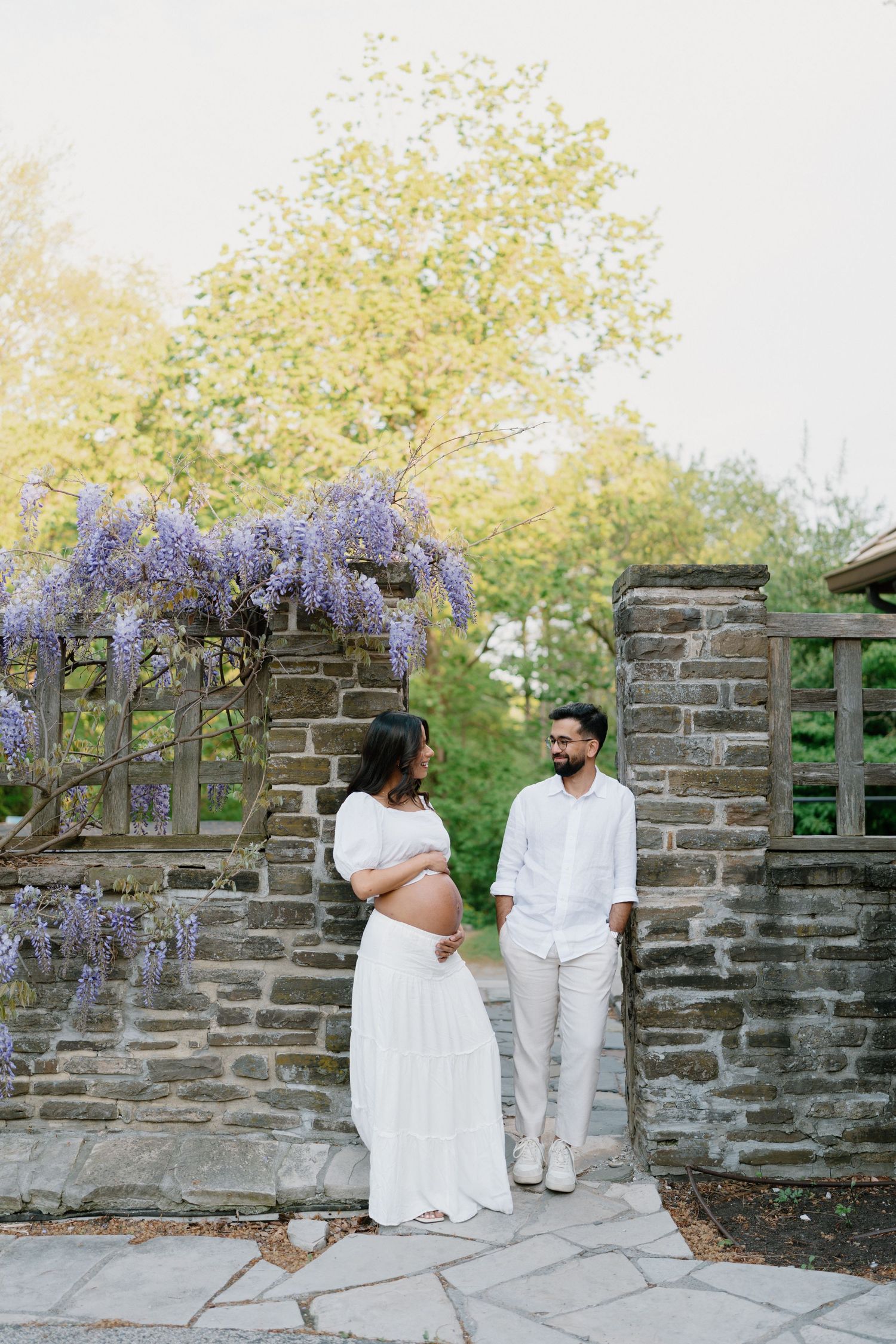 Pregnant woman and partner in white outfits standing before stone wall adorned with purple wisteria blooms.