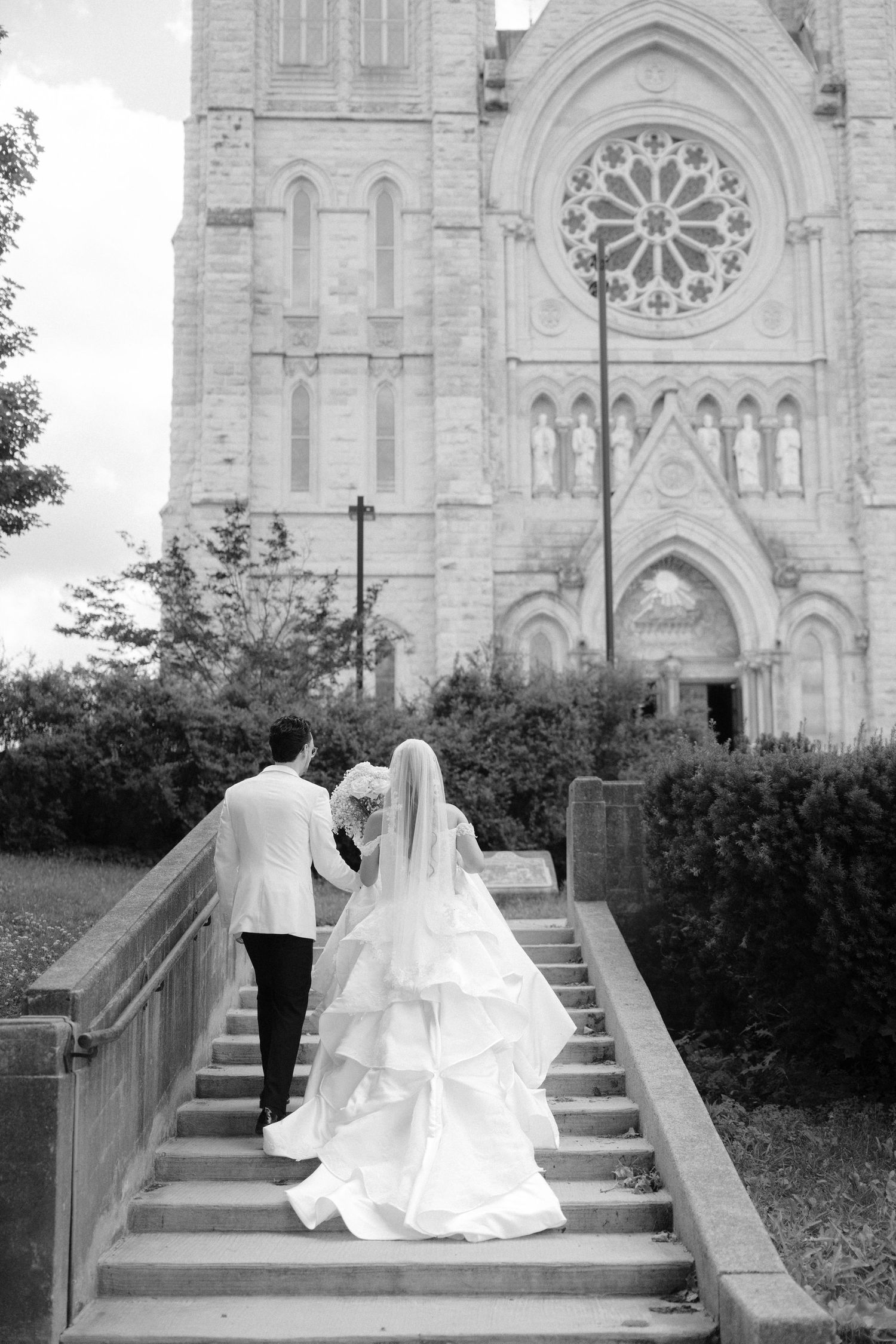 Black and white photo of bride and groom walking up stone steps toward a grand Gothic cathedral on their wedding day.