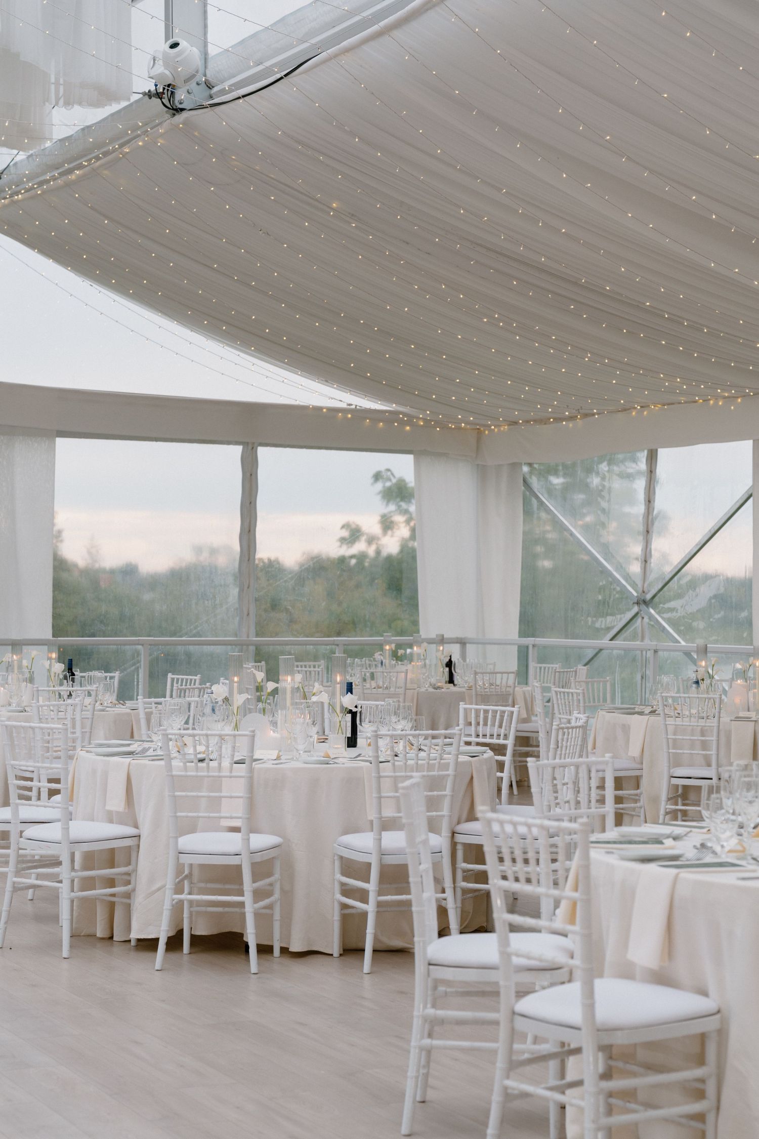 Elegant white wedding reception tent with fairy lights, chiavari chairs, and round tables overlooking lush greenery.