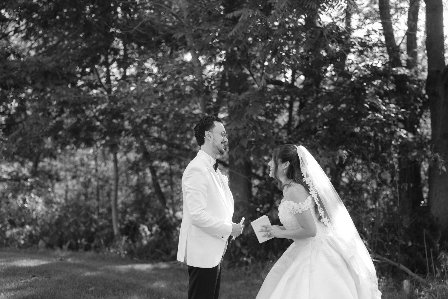 Black and white wedding photo of bride and groom sharing vows outdoors among lush trees.