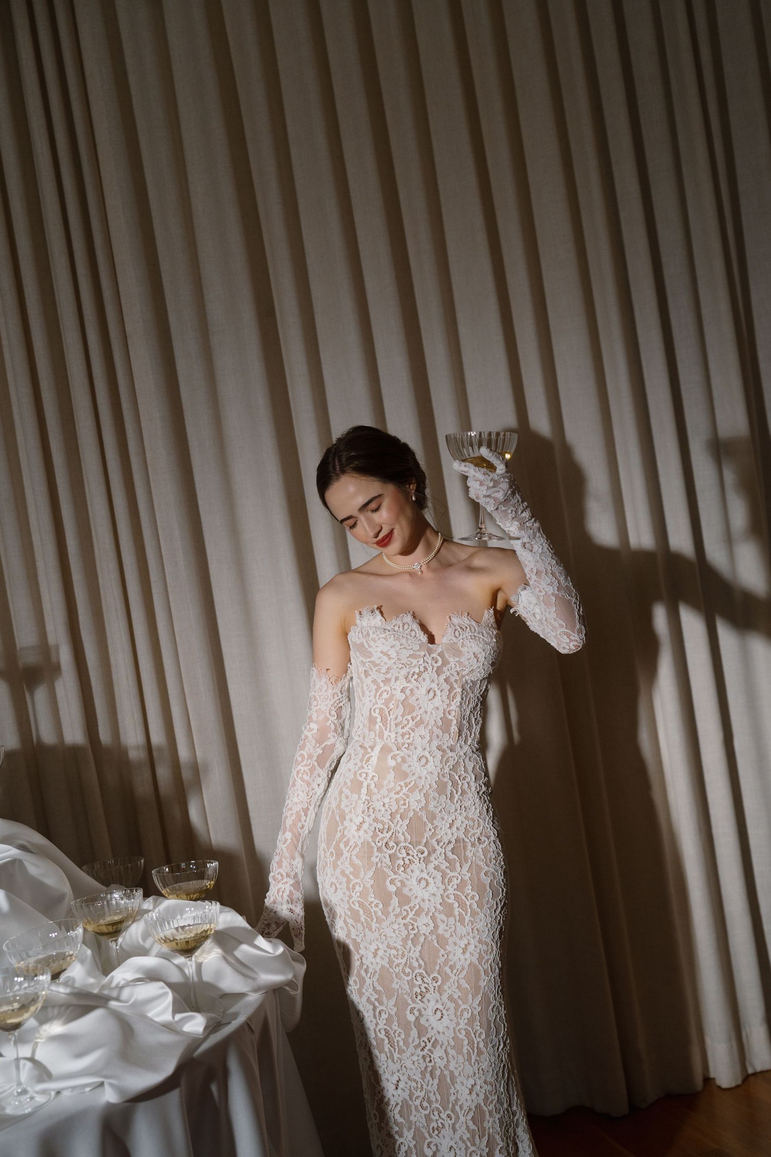 Elegant bride in lace strapless gown with long gloves holding champagne beside a wedding table.