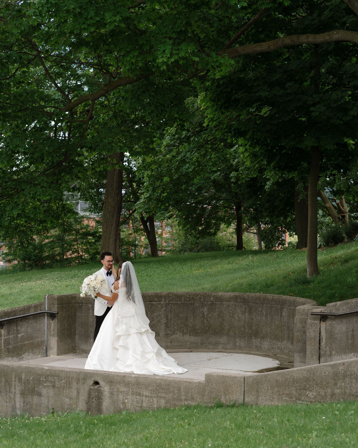Bride in white gown and groom in tuxedo pose in a stone amphitheater surrounded by lush green trees.