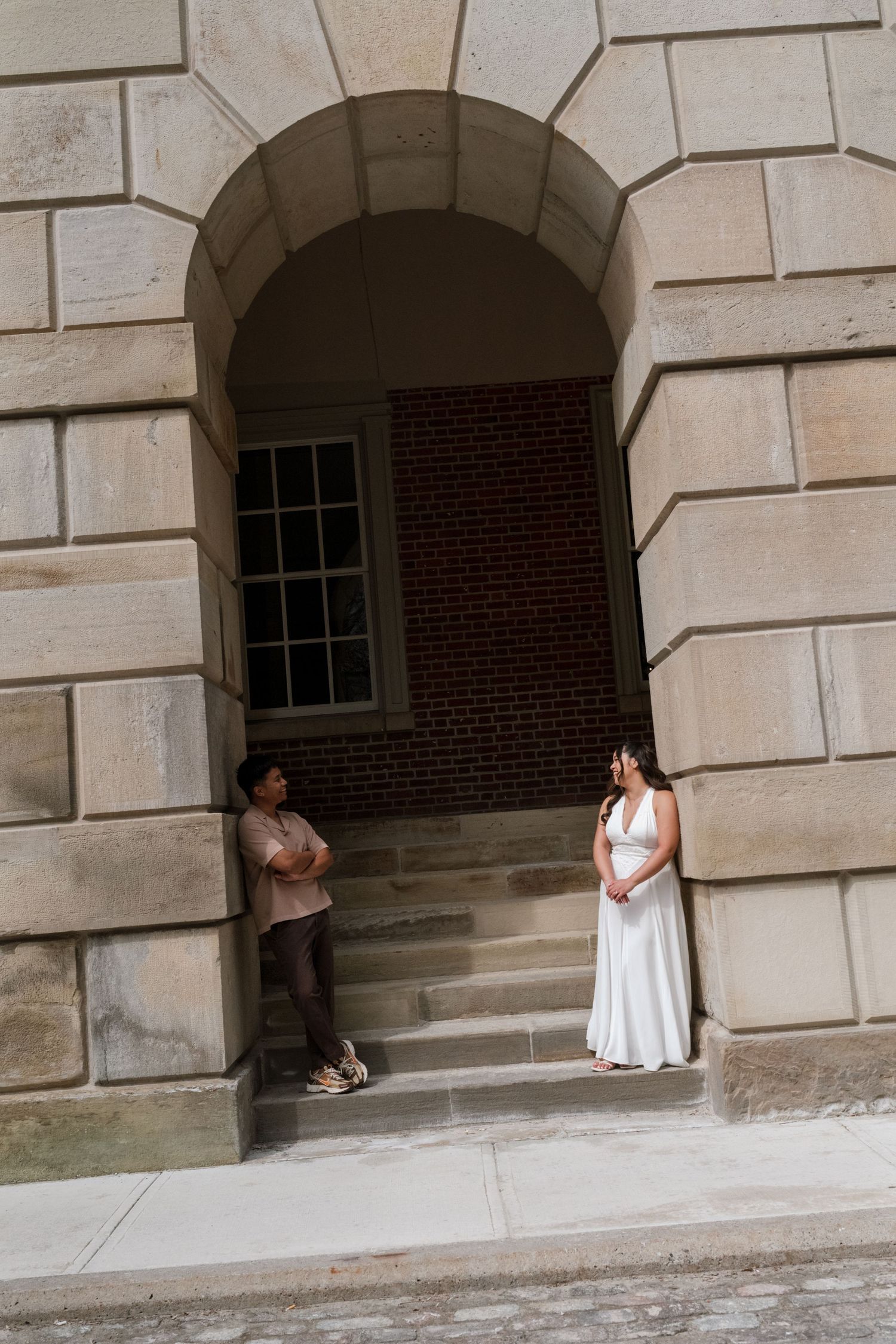 Bride in white gown and groom in casual attire stand apart under a stone archway of a historic building.