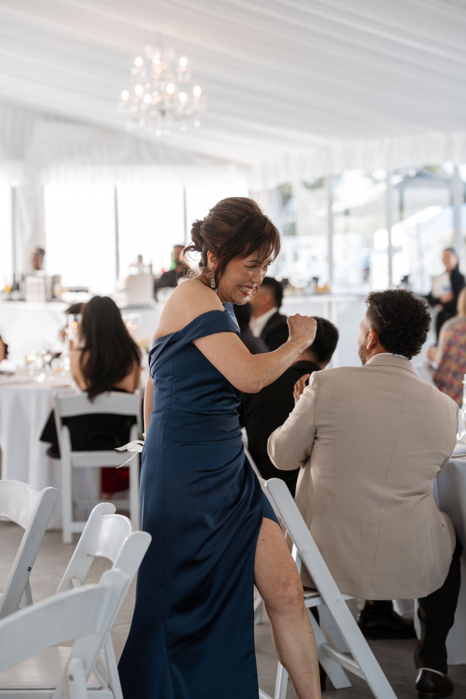 Wedding guests celebrate and dance during a reception in an elegant white tent venue with chandelier lighting.