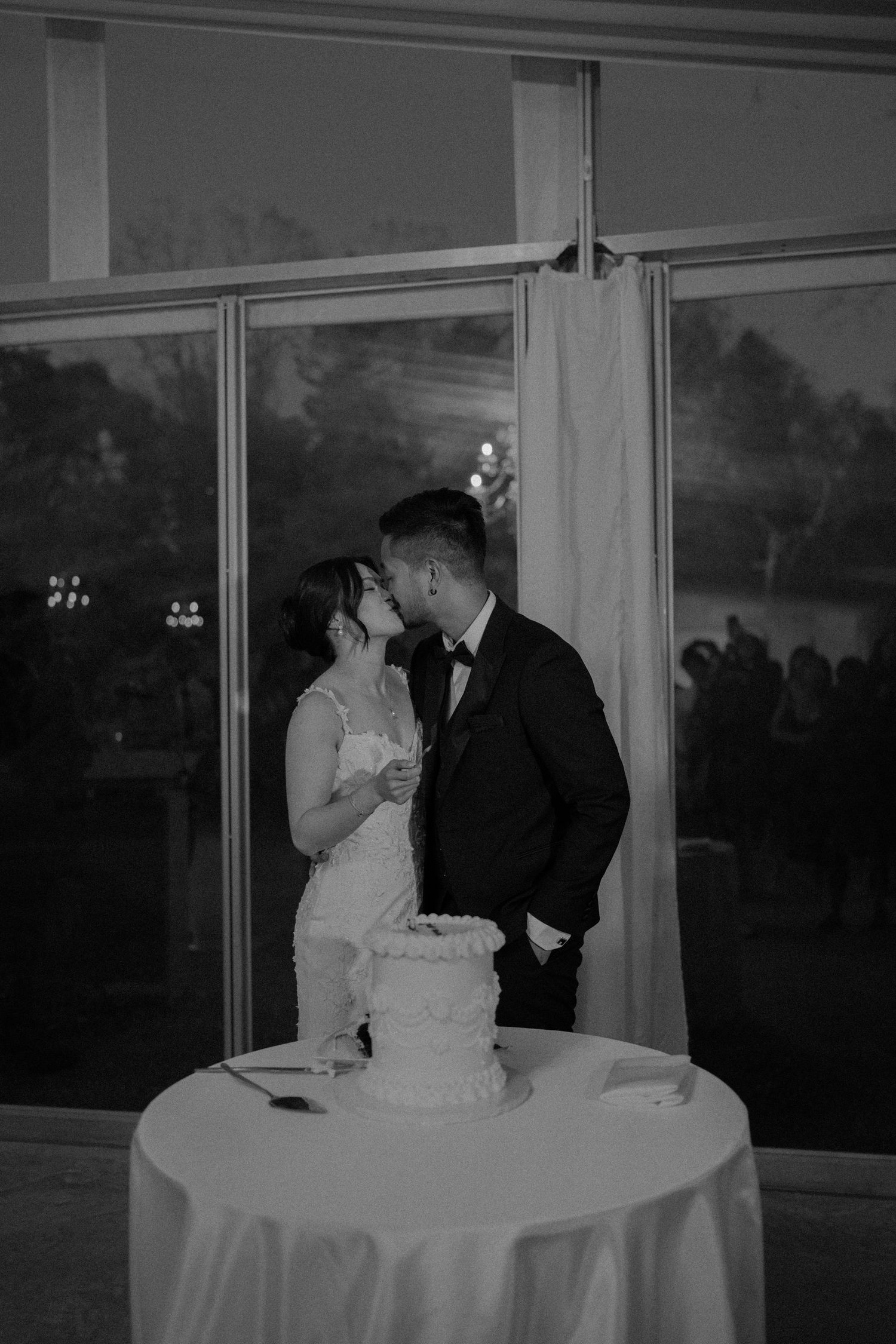 A couple shares a romantic moment at their wedding reception by a window with their white wedding cake in the foreground.