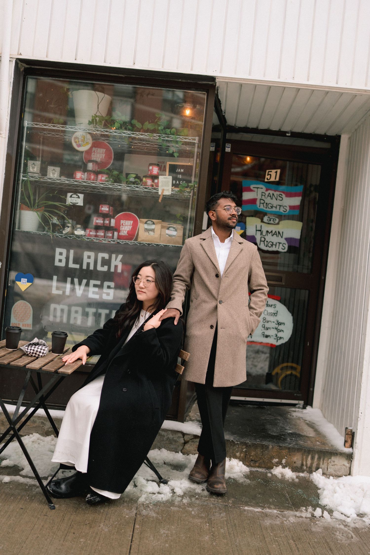 A couple in formal attire poses together in front of a storefront on a snowy winter day.