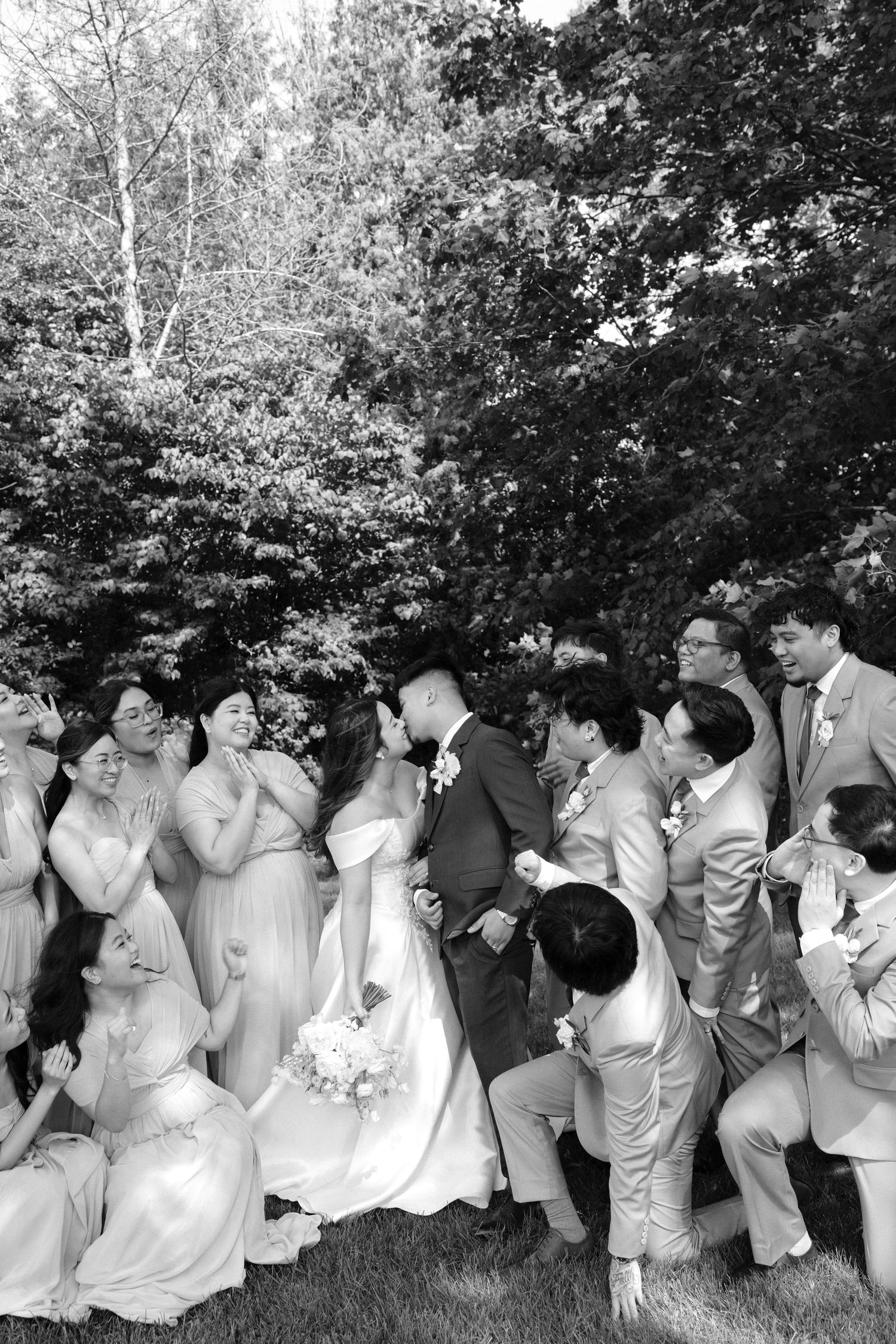 Black and white photo of a wedding party surrounding the bride and groom sharing a kiss outdoors.
