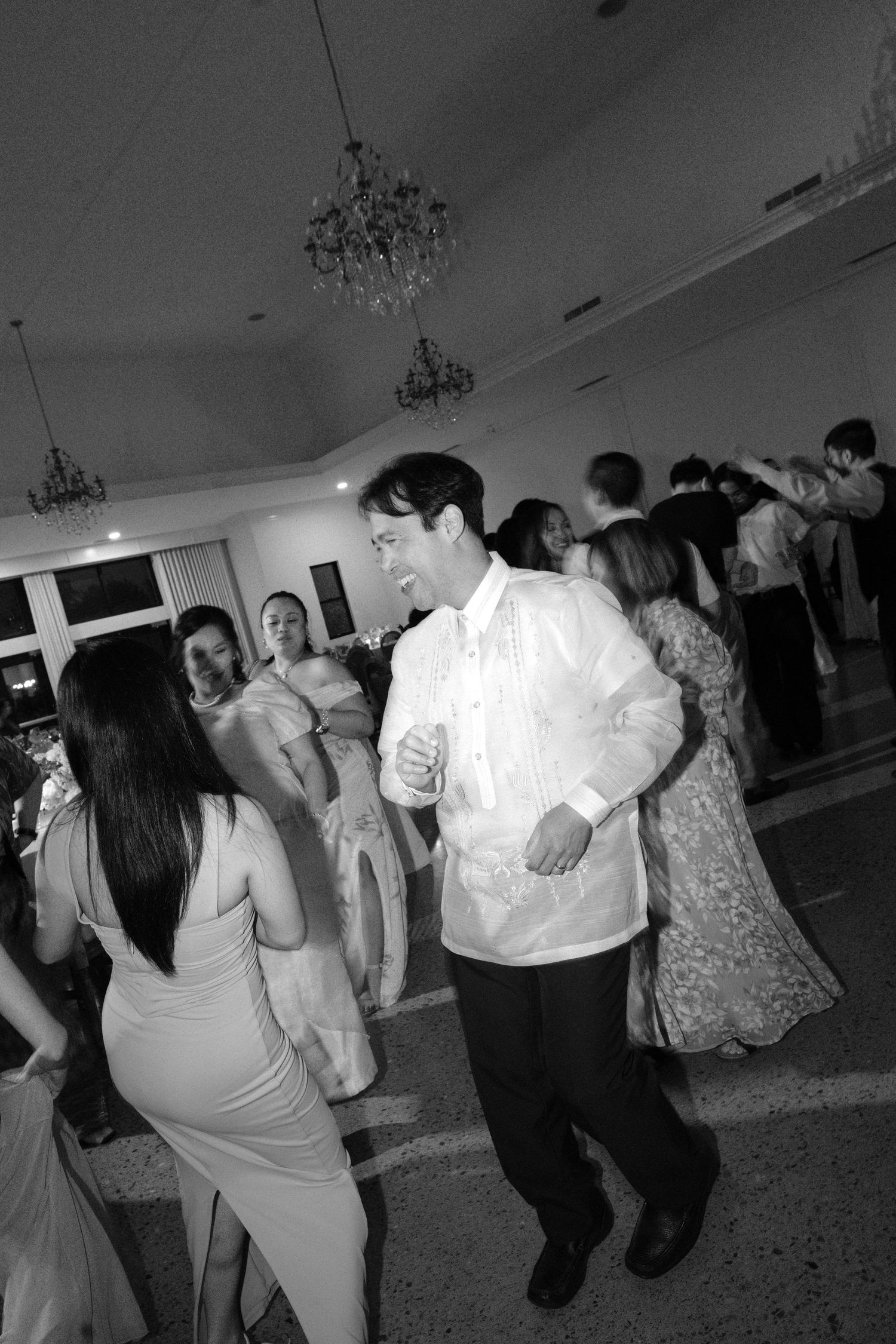 Black and white photo of elegantly dressed guests dancing at a formal wedding reception in a chandelier-lit ballroom.