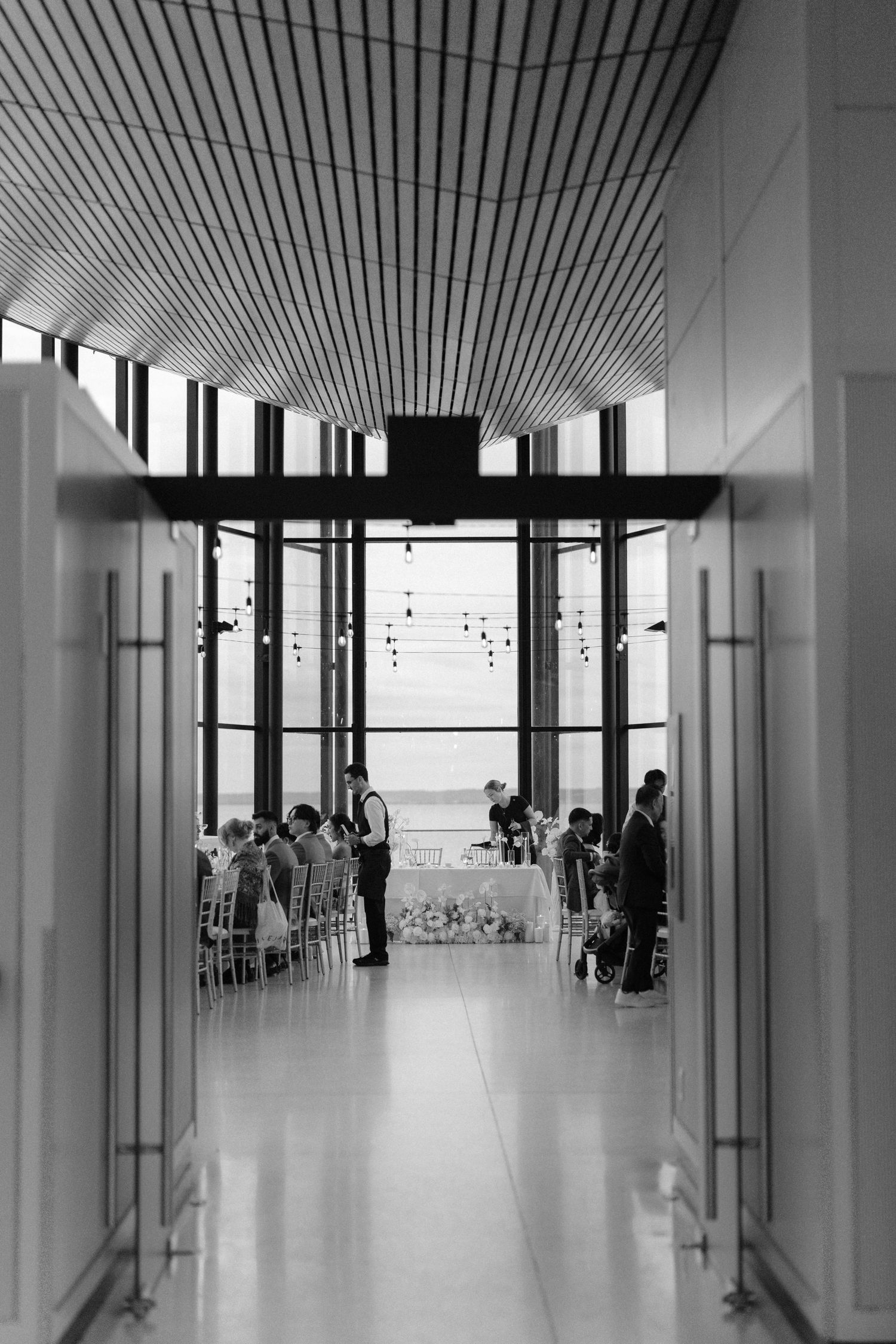 Black and white photo of an elegant wedding ceremony in a modern venue with floor-to-ceiling windows and string lights.