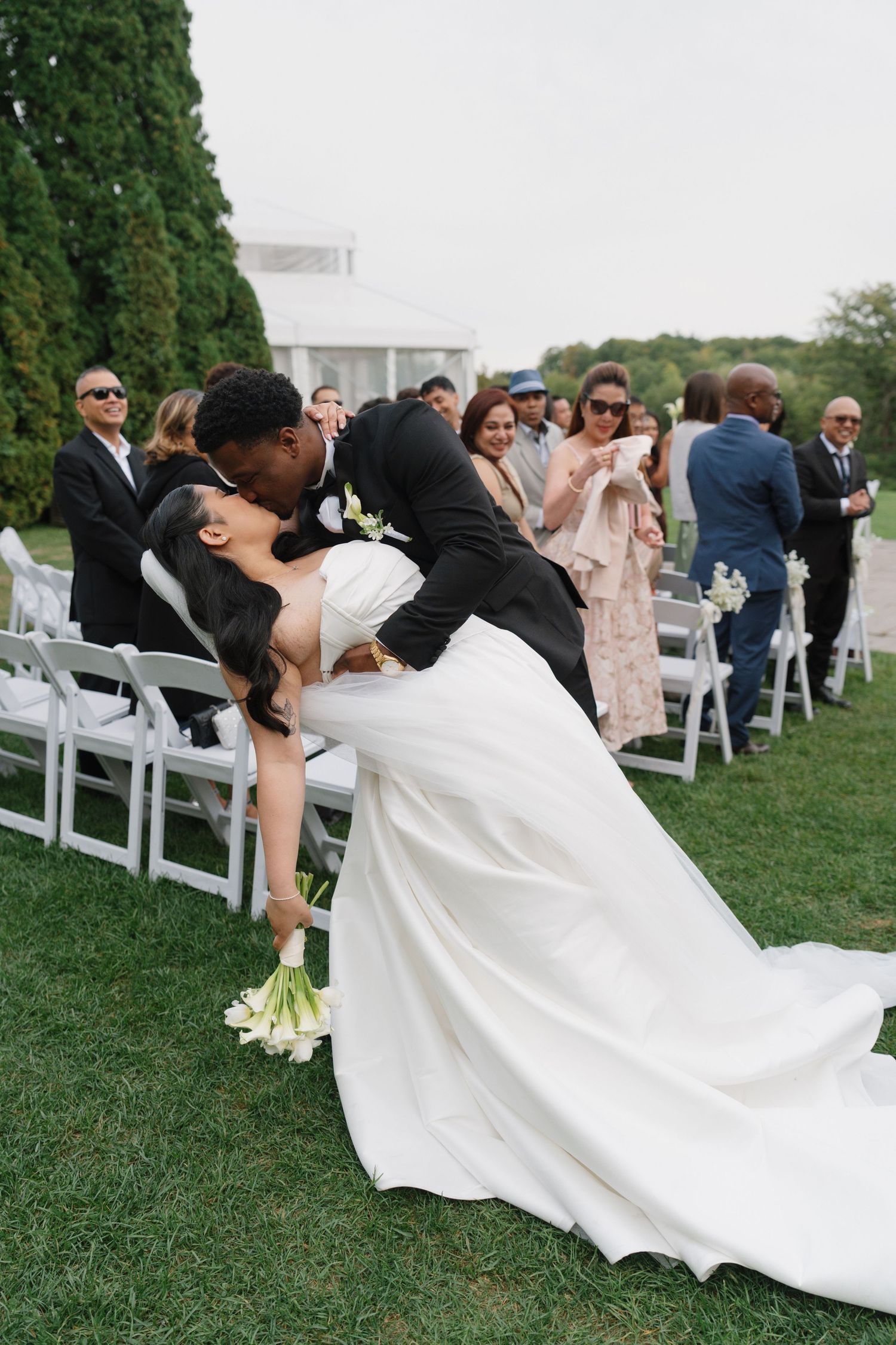 Groom dips bride for a romantic kiss at outdoor wedding ceremony on green lawn with guests watching.