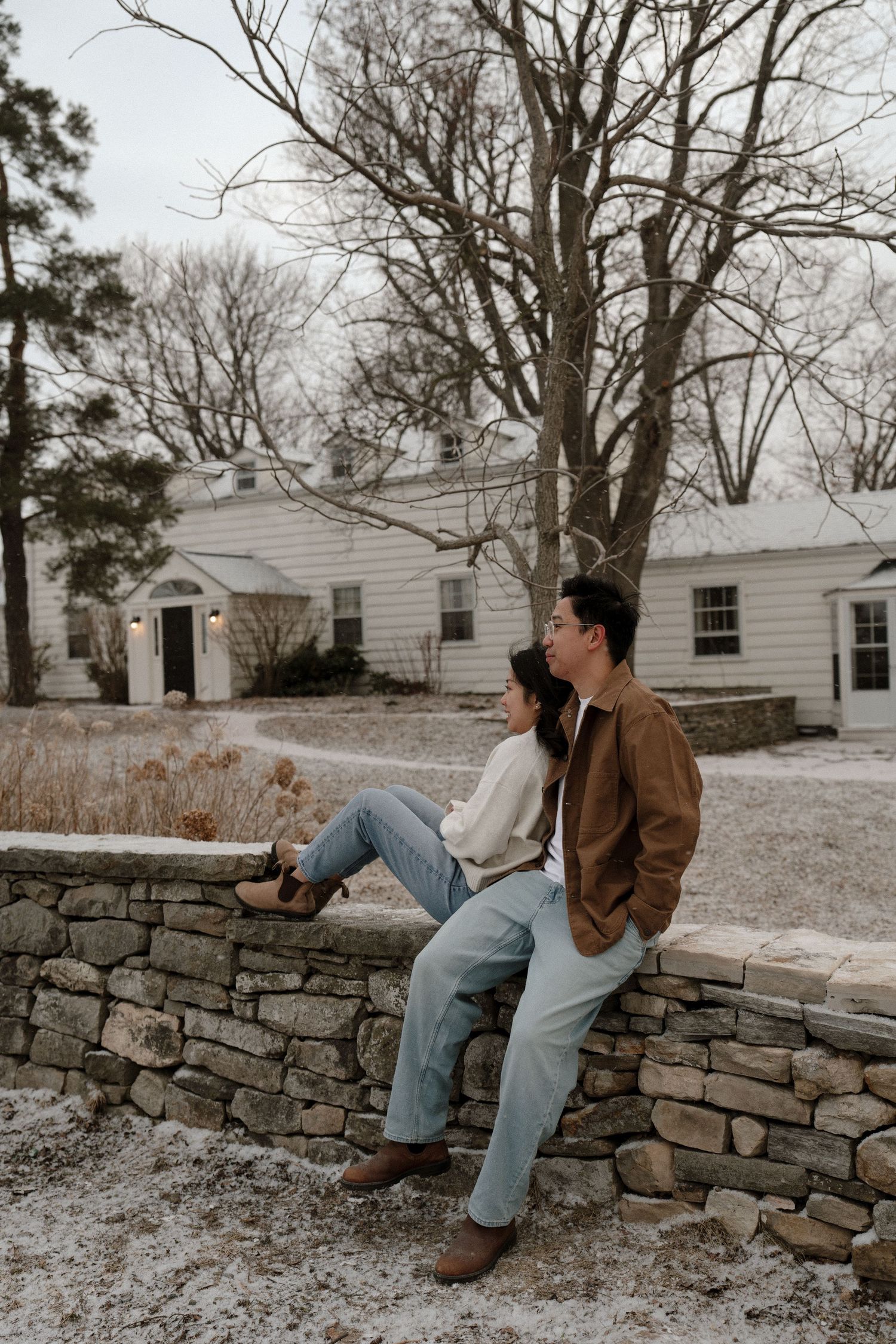 Couple sits together on a stone wall in winter landscape, wearing casual earth-tone outfits.