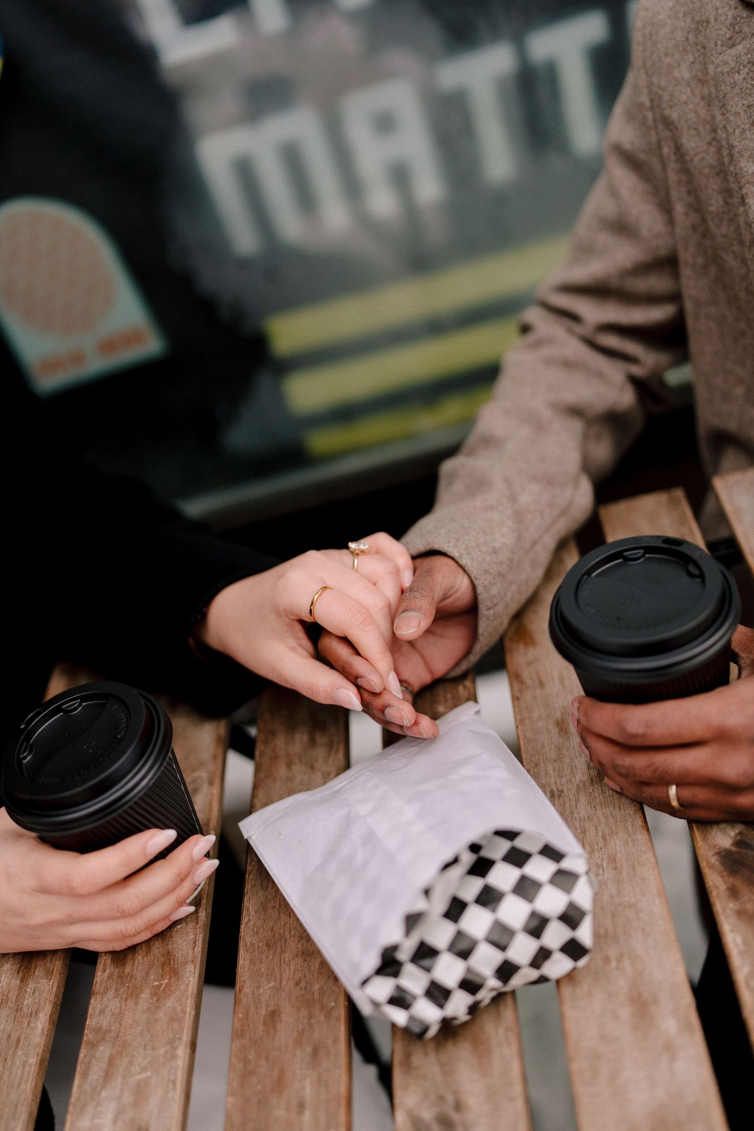 Two hands hold coffee cups and a napkin with checkered pattern against a wooden fence background.