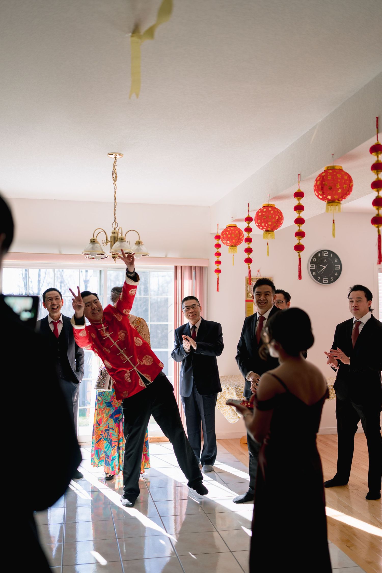 Groom in red traditional Chinese wedding attire celebrates with guests in a decorated room with red lanterns.