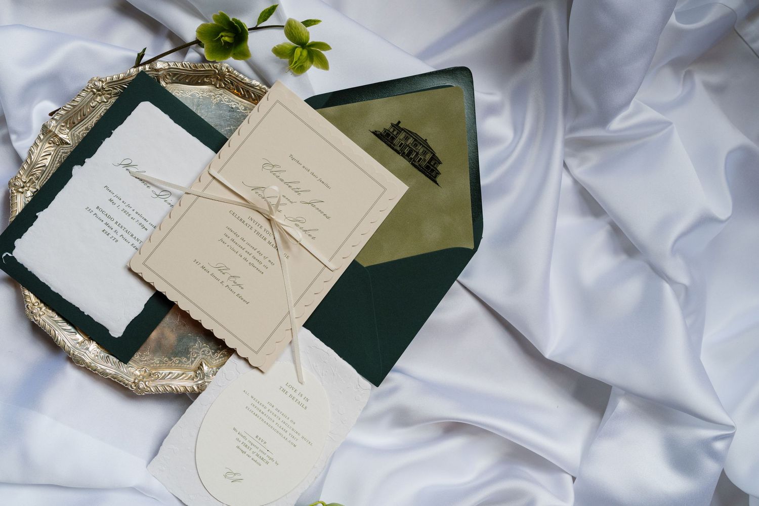 Elegant wedding invitation suite with dark green envelopes and cream cards on white satin fabric.