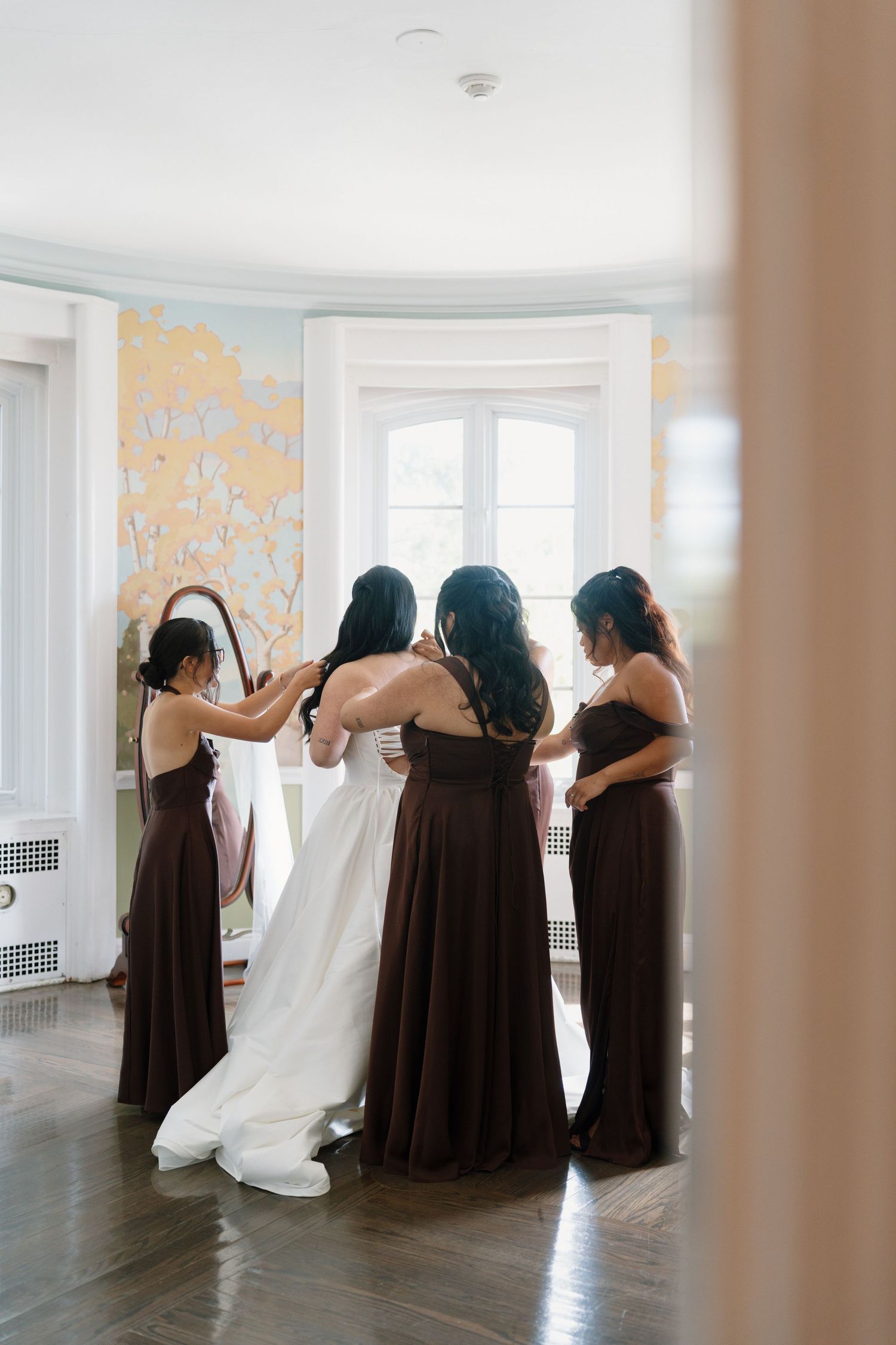 Bridesmaids in dark brown gowns help bride in white wedding dress prepare in elegant room with floral mural wallpaper.