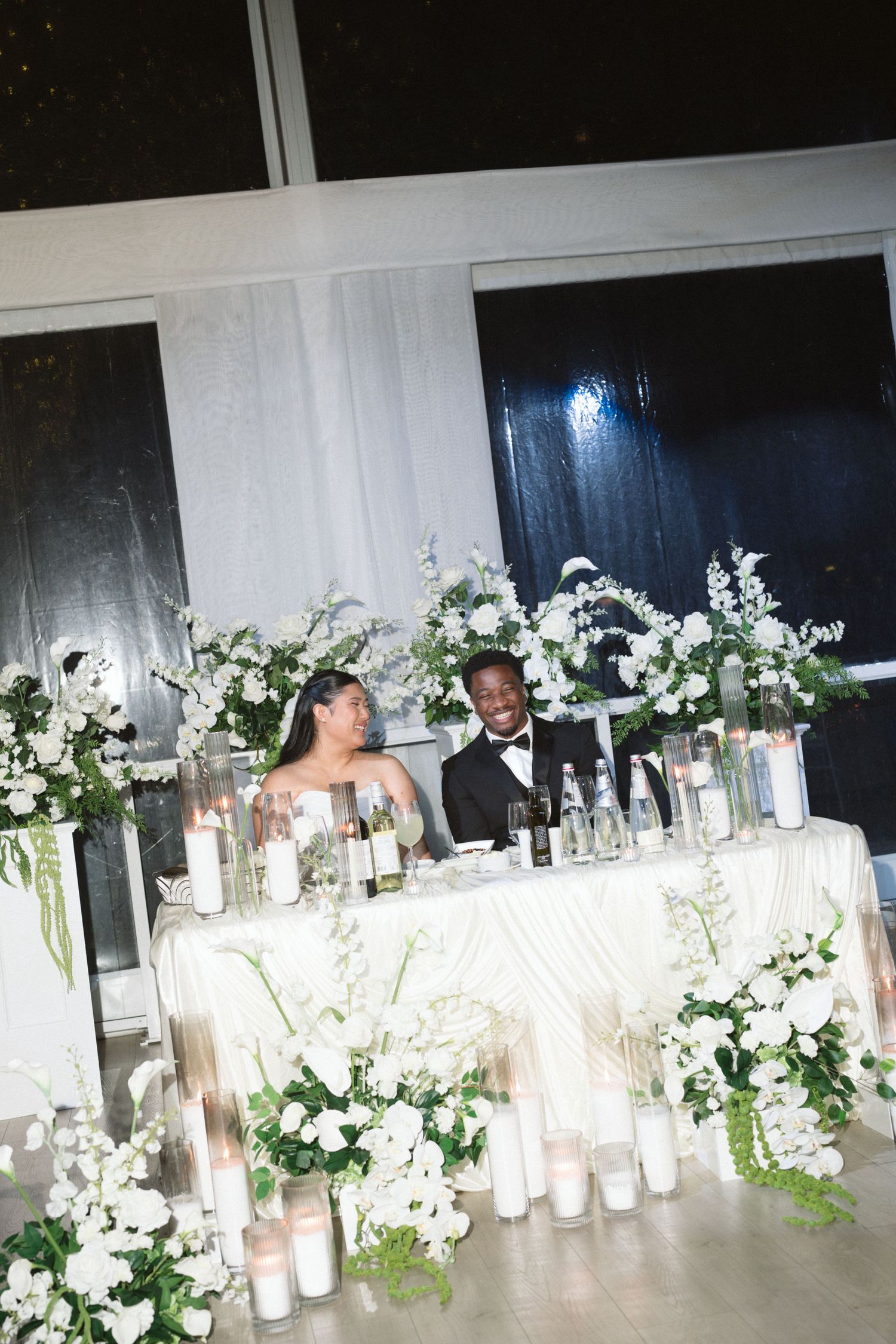 Bride and groom seated at sweetheart table adorned with white floral arrangements and candles at elegant wedding reception.