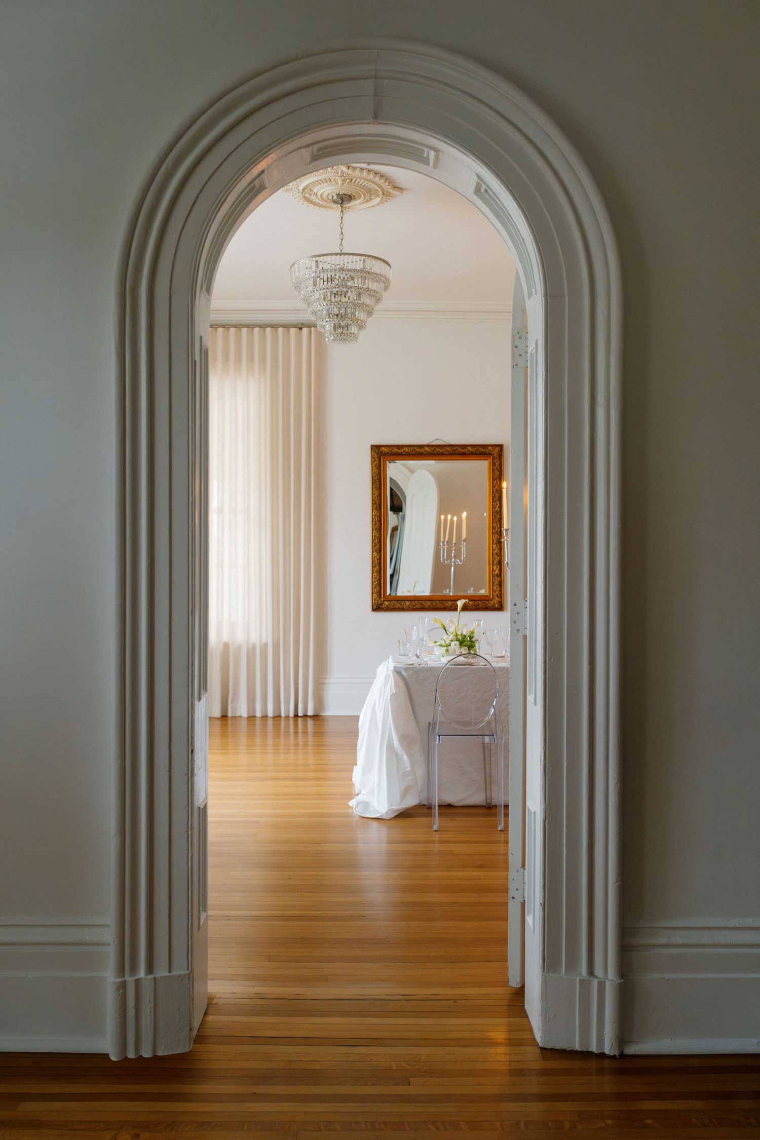 Elegant white archway leading to a bright wedding reception room with chandelier and gold mirror.