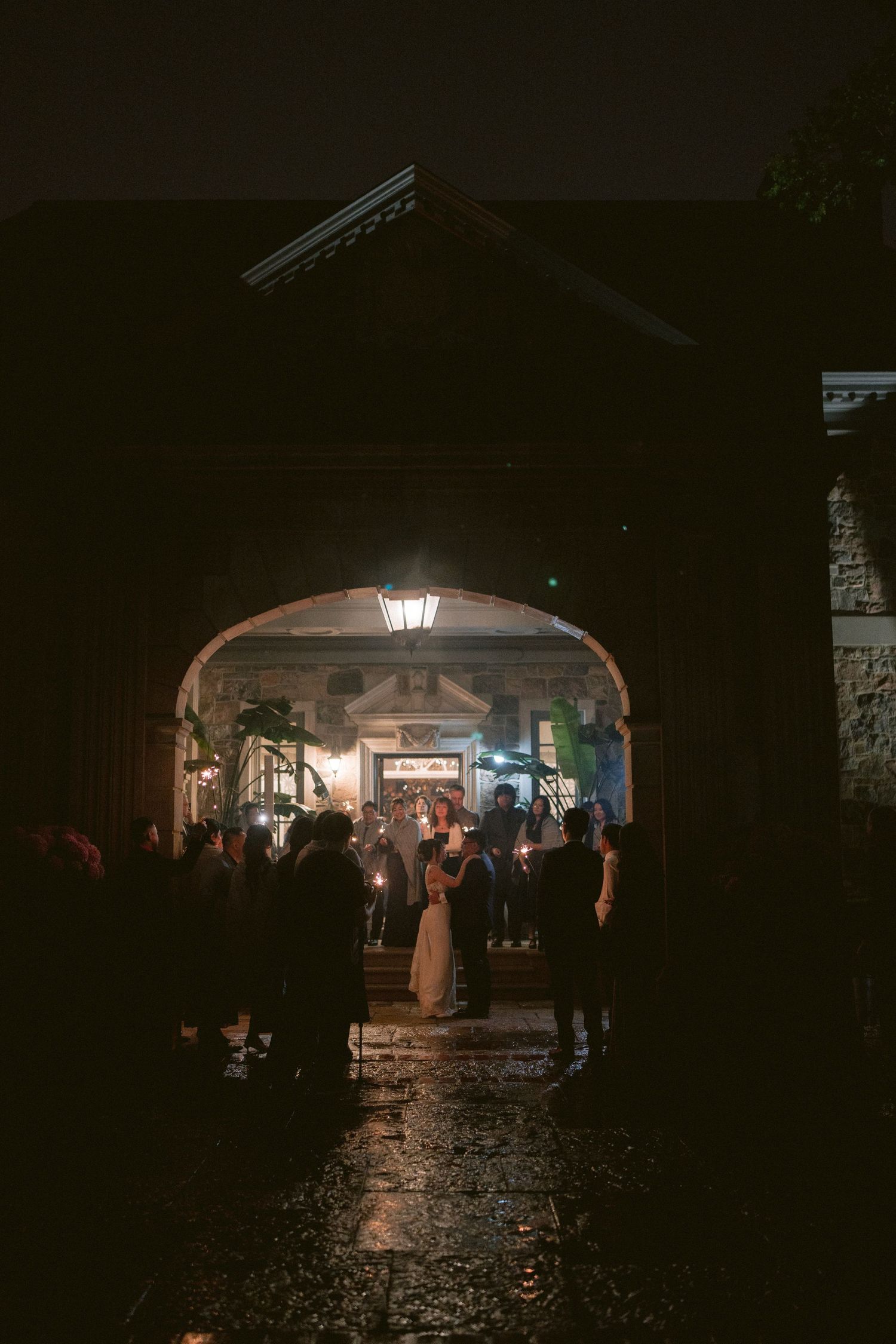 Wedding guests gather under a stone archway at night, illuminated by lantern light on wet cobblestones.