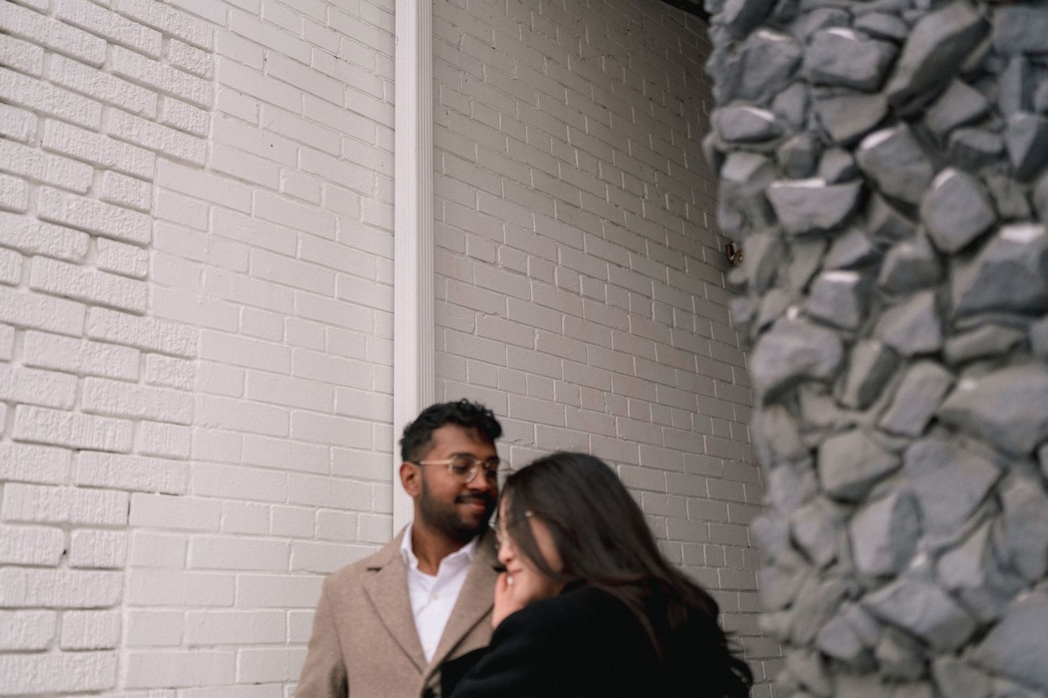 A romantic couple shares an intimate moment against a white brick wall and stone column.