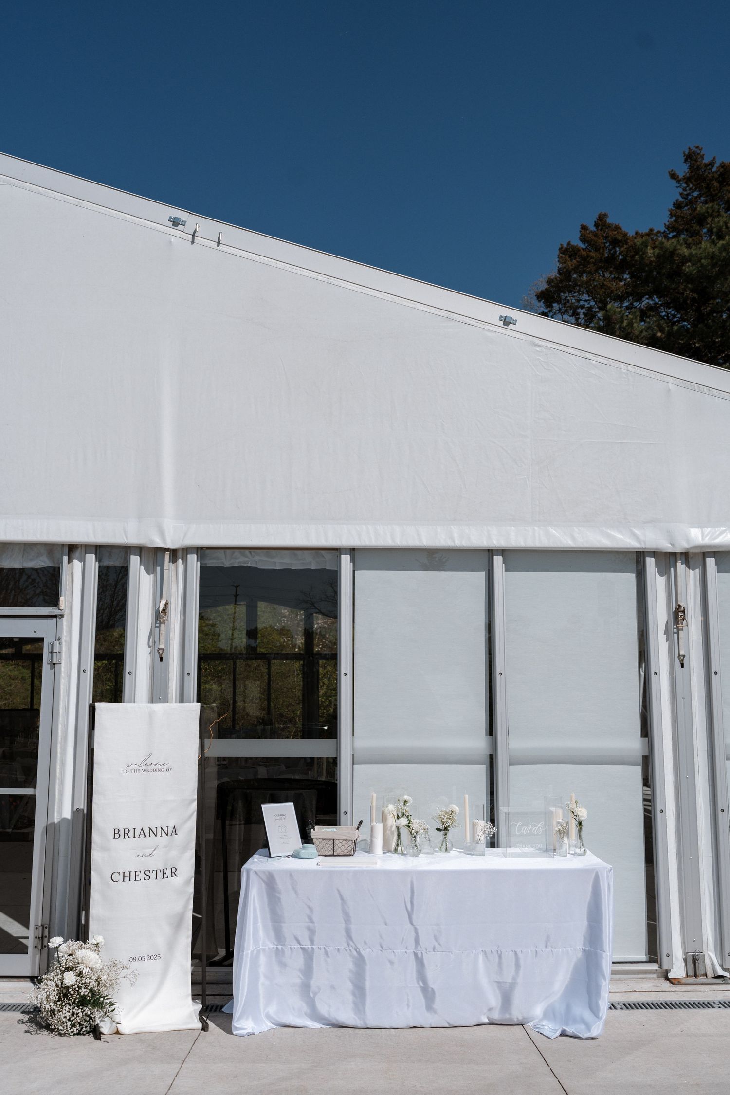 White wedding reception tent with welcome table and floral arrangements under a bright blue sky.