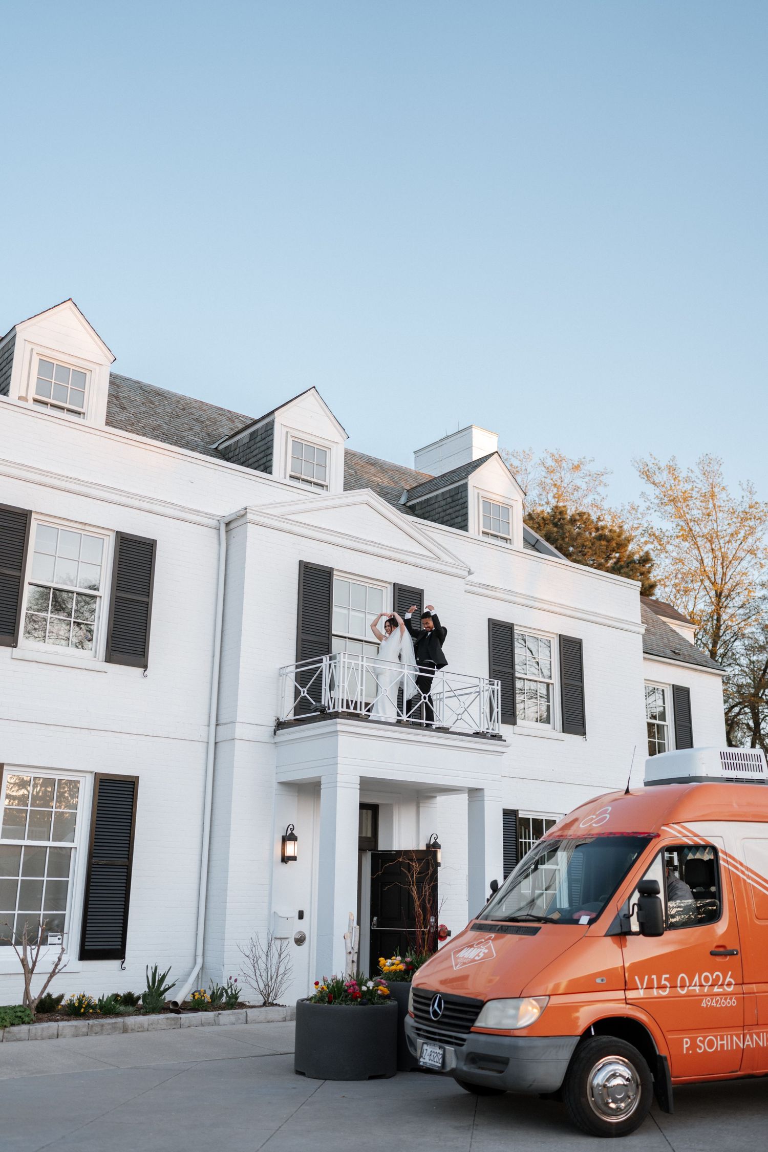 A white colonial style house with dormers and a balcony, with an orange delivery van parked in front.