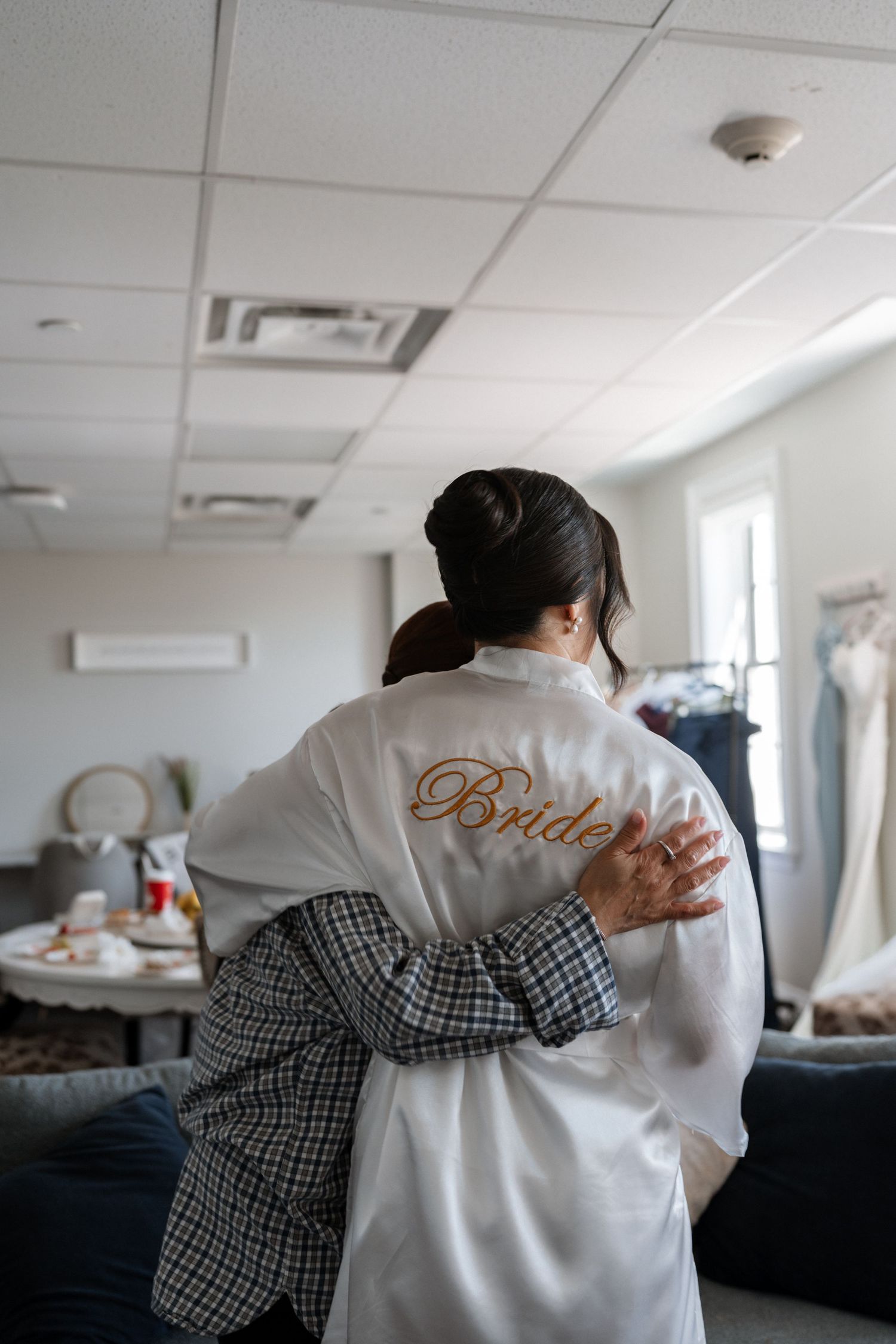 A person wearing a white satin robe with 'Bride' embroidery is hugged from behind in a bridal preparation room.