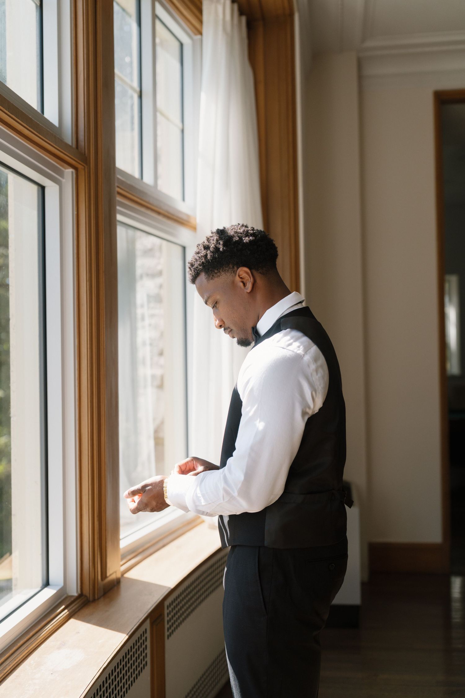 Elegant groom in white shirt and black vest adjusting cufflinks by a sunlit window before wedding ceremony.