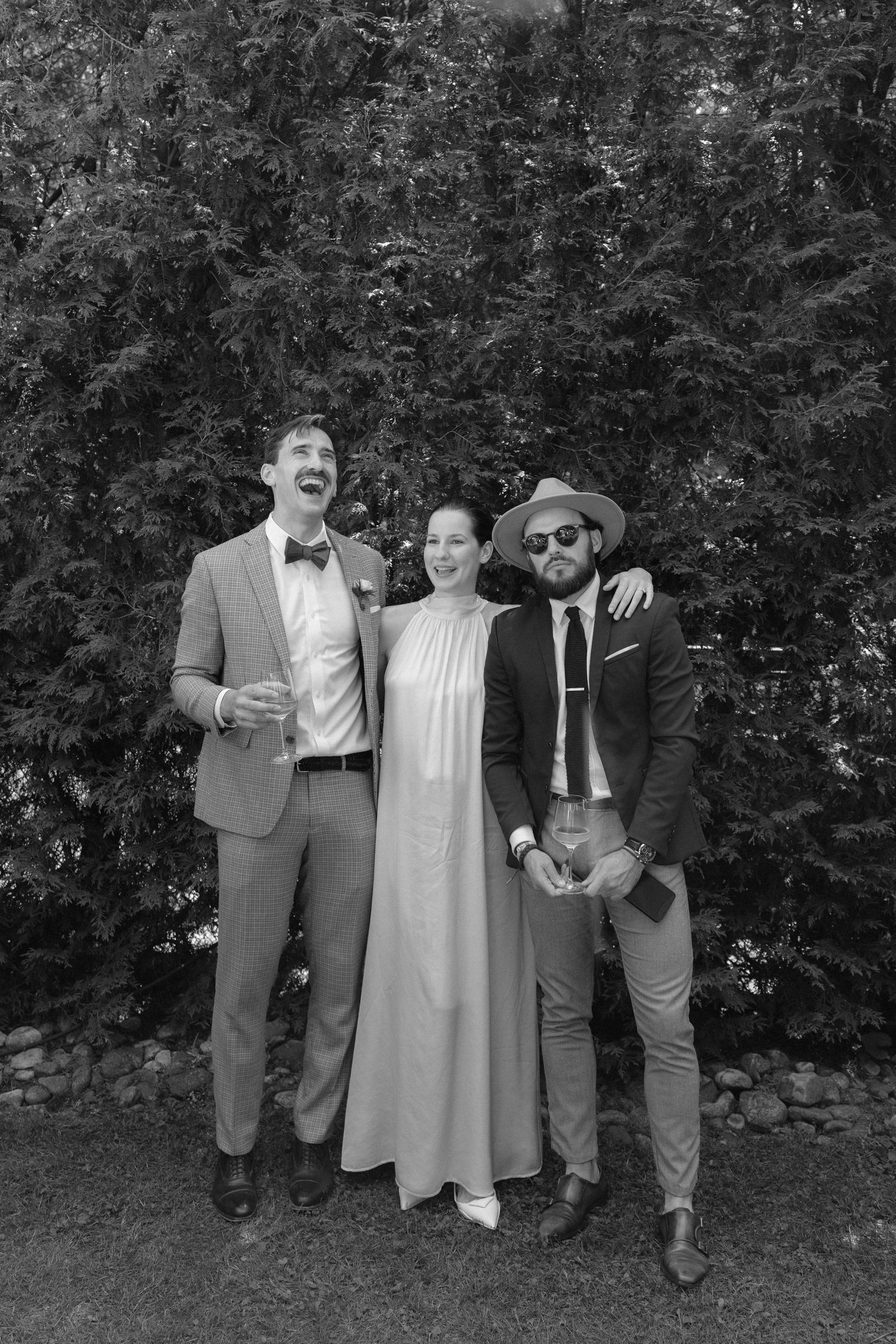 Black and white photo of two men and a woman posing outdoors at a wedding, with evergreen trees in background.