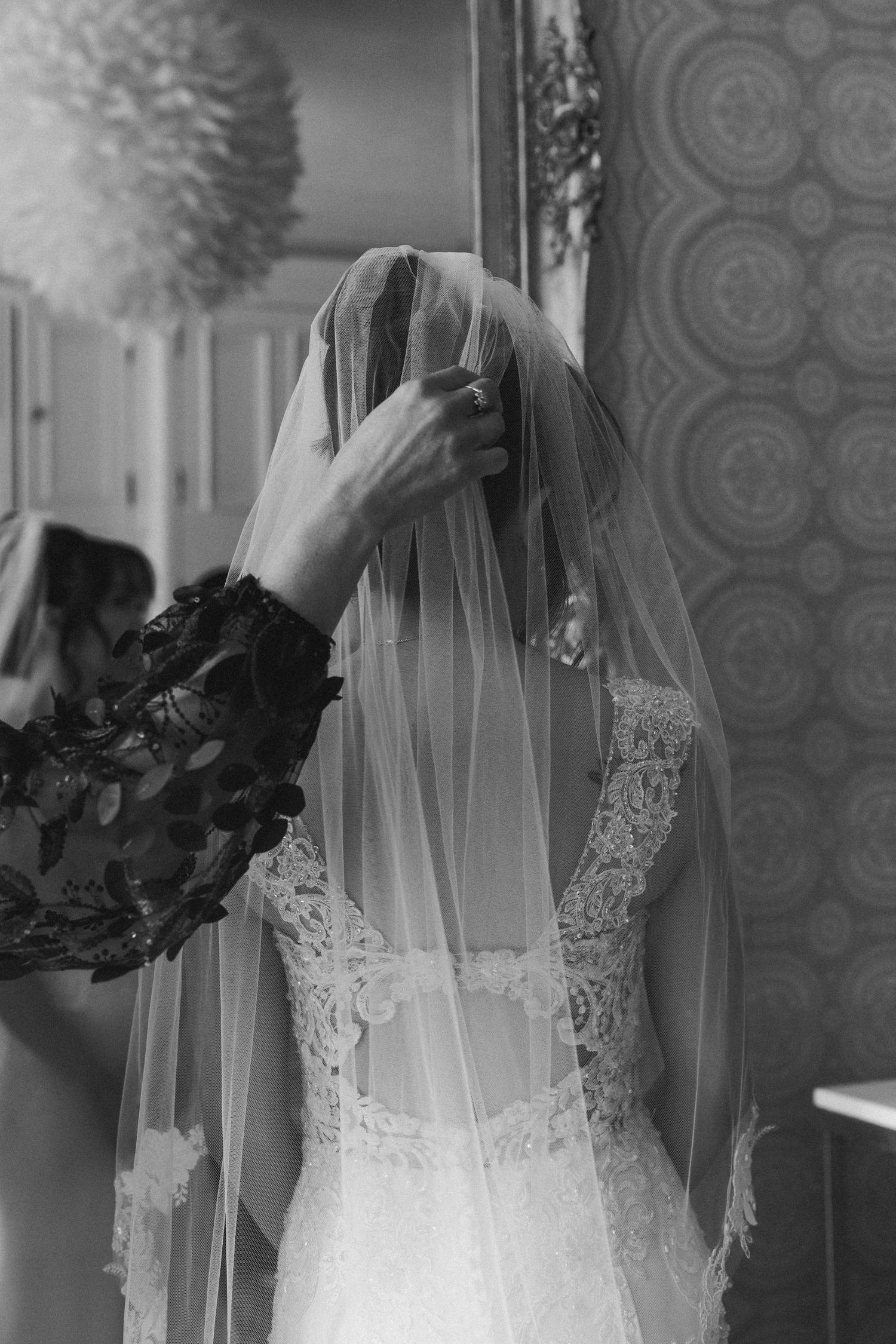 Black and white photo of hands adjusting a delicate bridal veil against an ornate wallpaper backdrop.