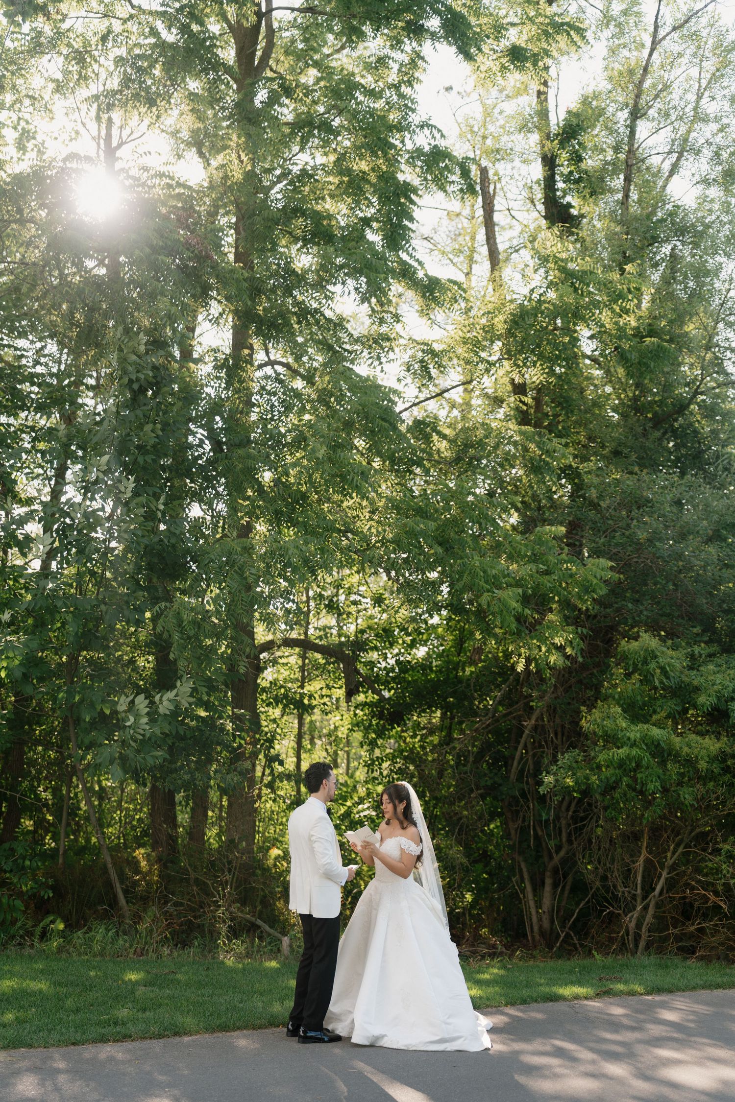 Bride and groom share a romantic first look moment on a garden path surrounded by lush green trees with sunlight filtering through.