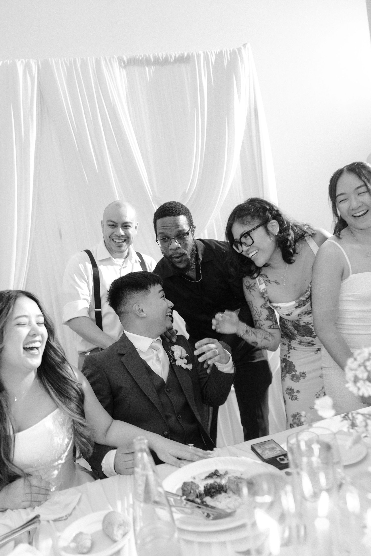 Black and white photo of wedding guests laughing and celebrating together at a reception dinner table.