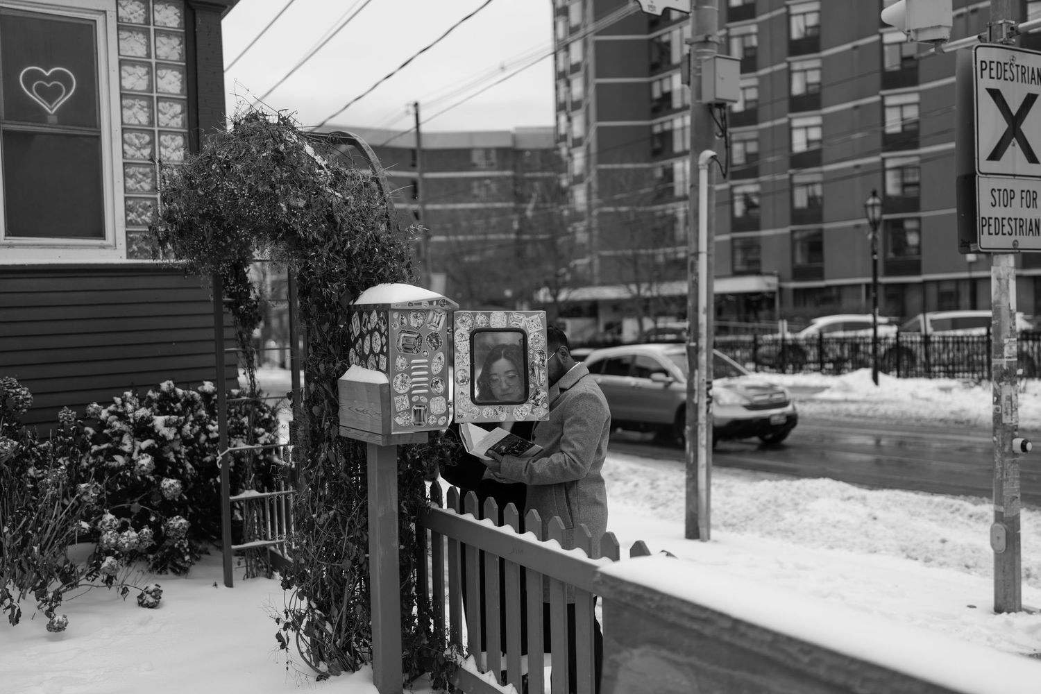 A black and white winter scene showing a snow-covered fence and garden with apartment buildings in the background.