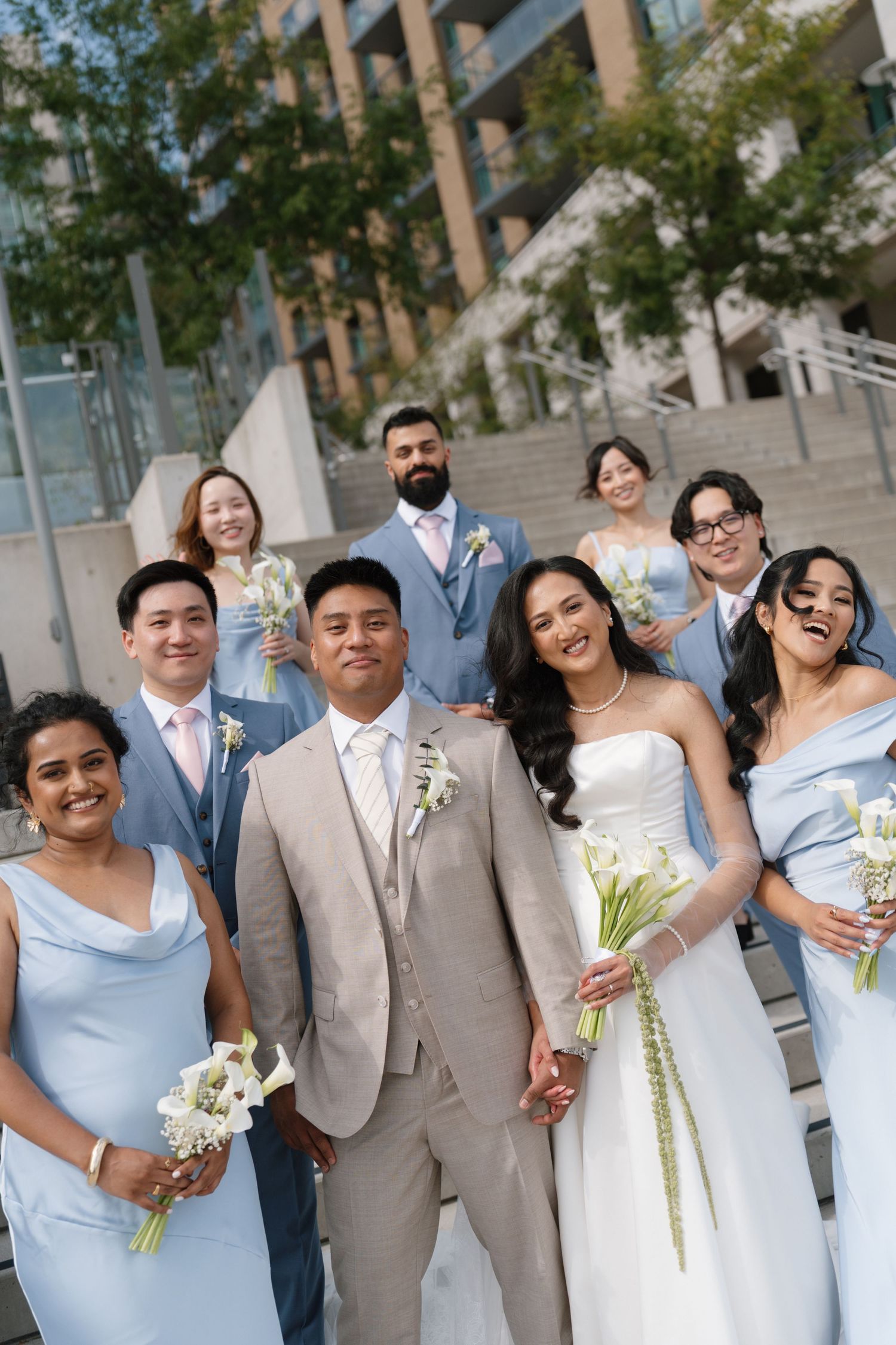 Diverse wedding party poses outdoors in blue bridesmaid dresses and tan suit, bride holds white calla lilies bouquet.