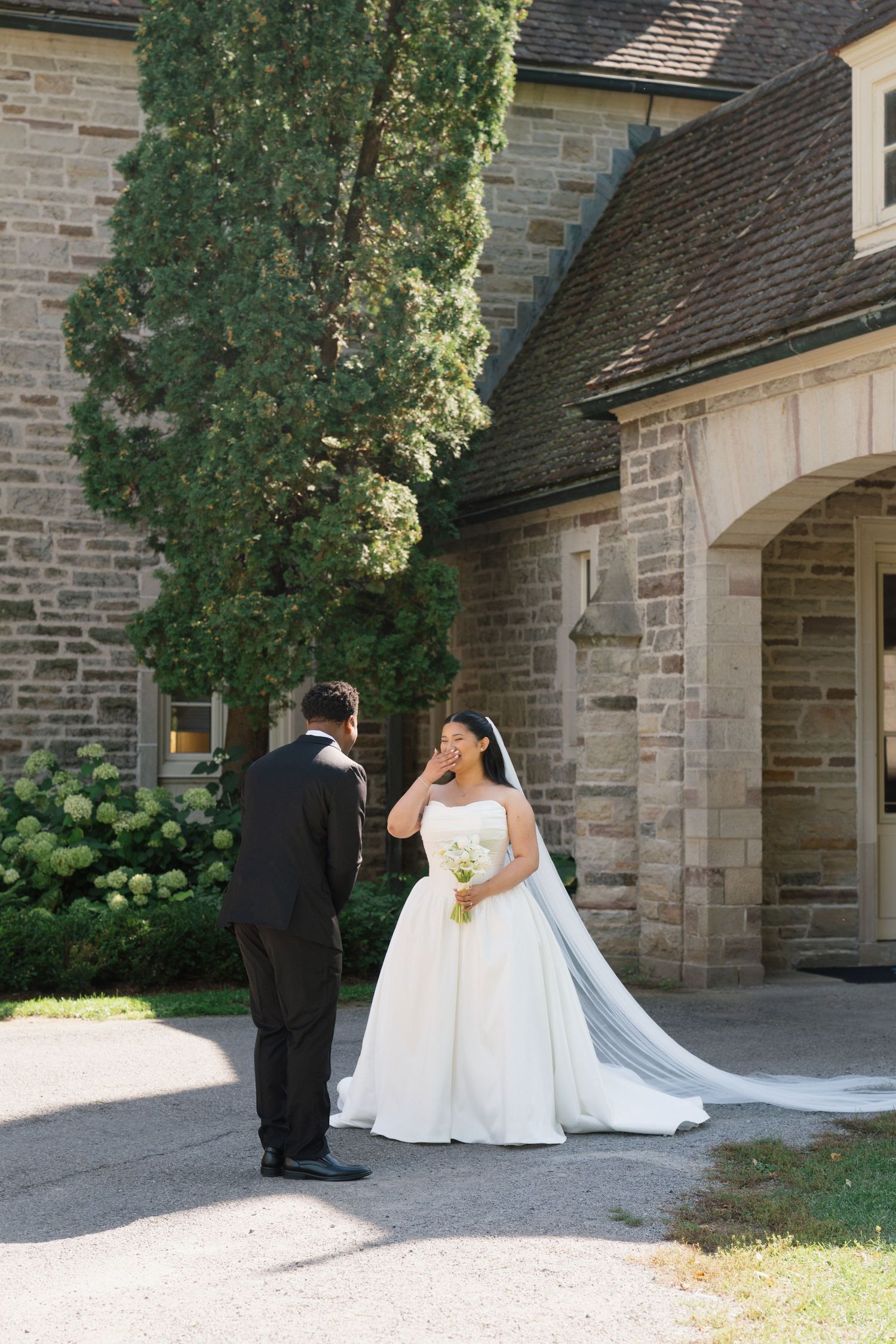 Bride in white gown holds bouquet facing groom in tuxedo outside stone church during outdoor wedding first look.