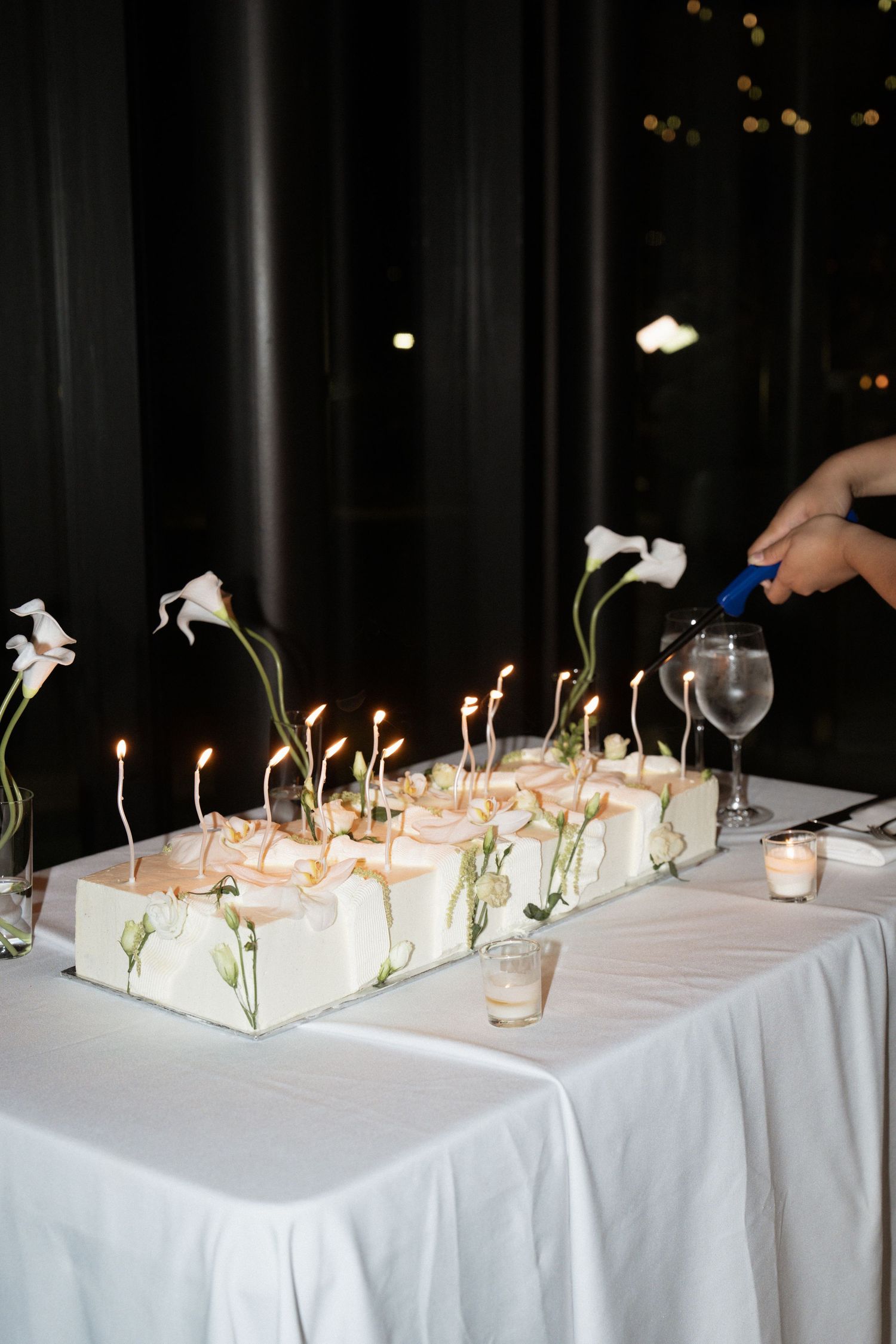 Elegant white wedding cake with lit candles and calla lily flowers on a white tablecloth, being lit at a reception.