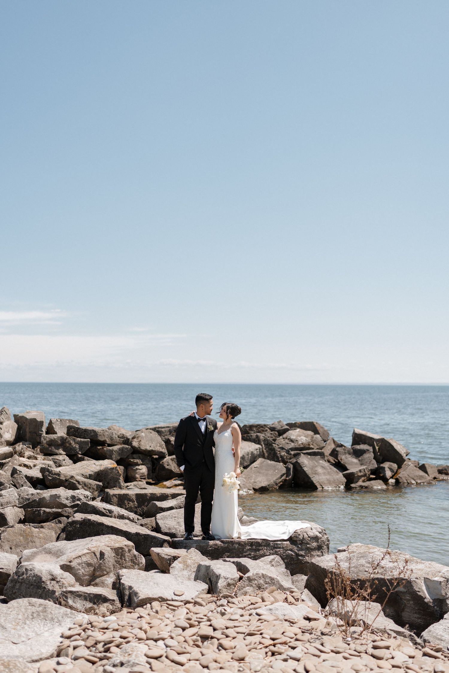 A couple in wedding attire stands together on rocky waterfront overlooking a serene ocean under bright blue skies.