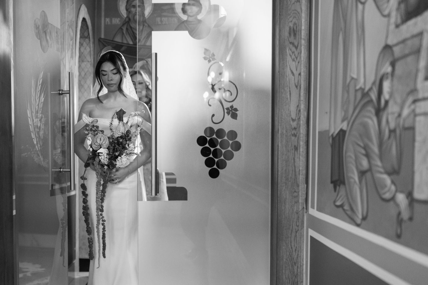 Black and white photo of a bride in a white gown holding a bouquet, standing in a doorway with decorative wall art.