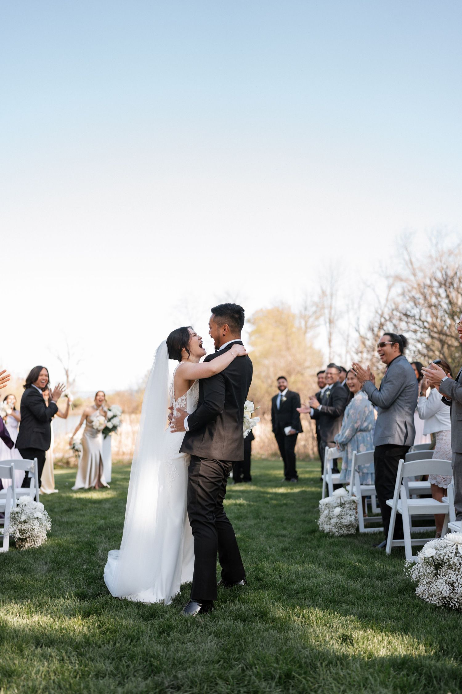 Wedding guests watching newlyweds share their first dance on a sunlit lawn with white chairs.