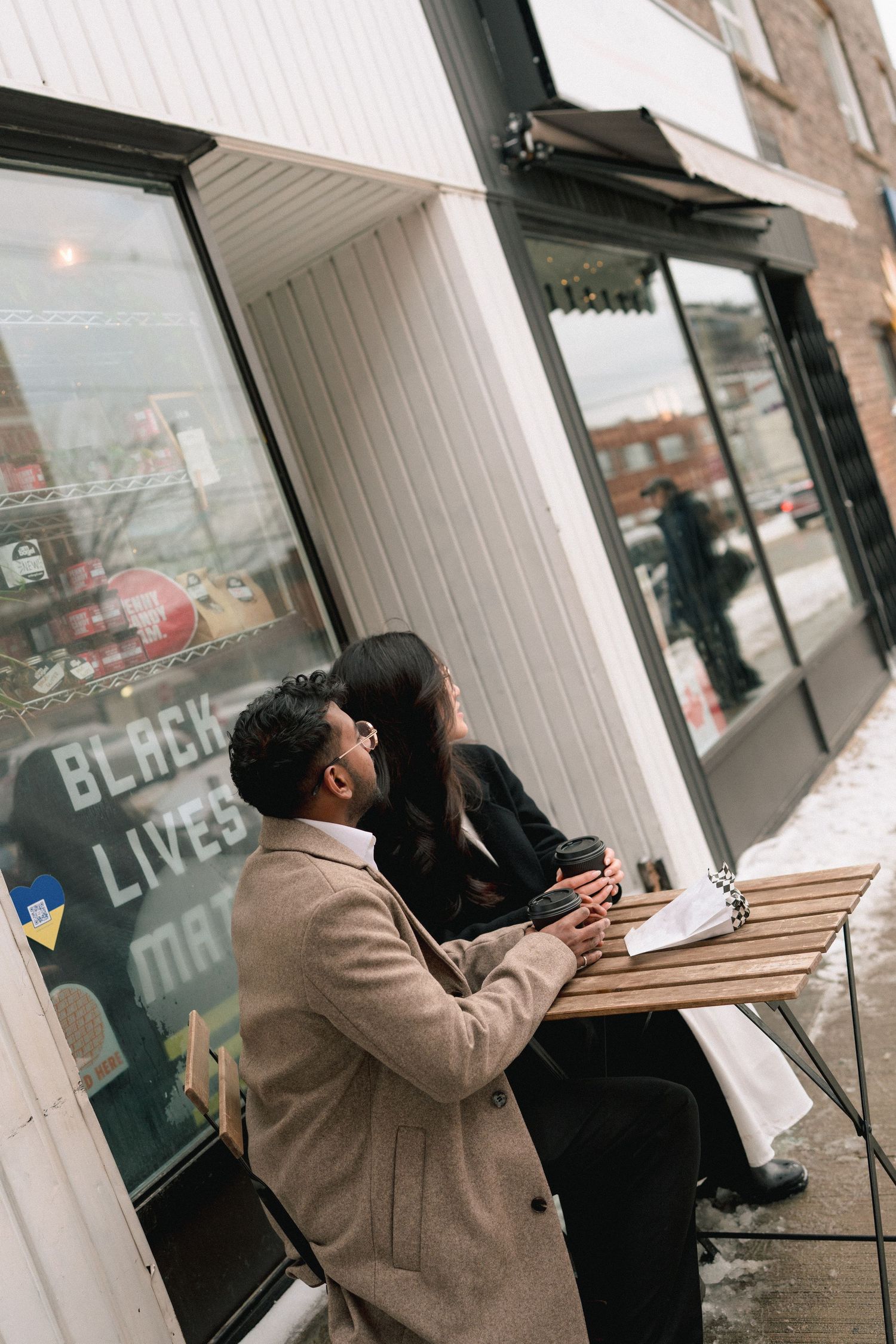 Two people sit at an outdoor cafe table having coffee and engaging in animated conversation.