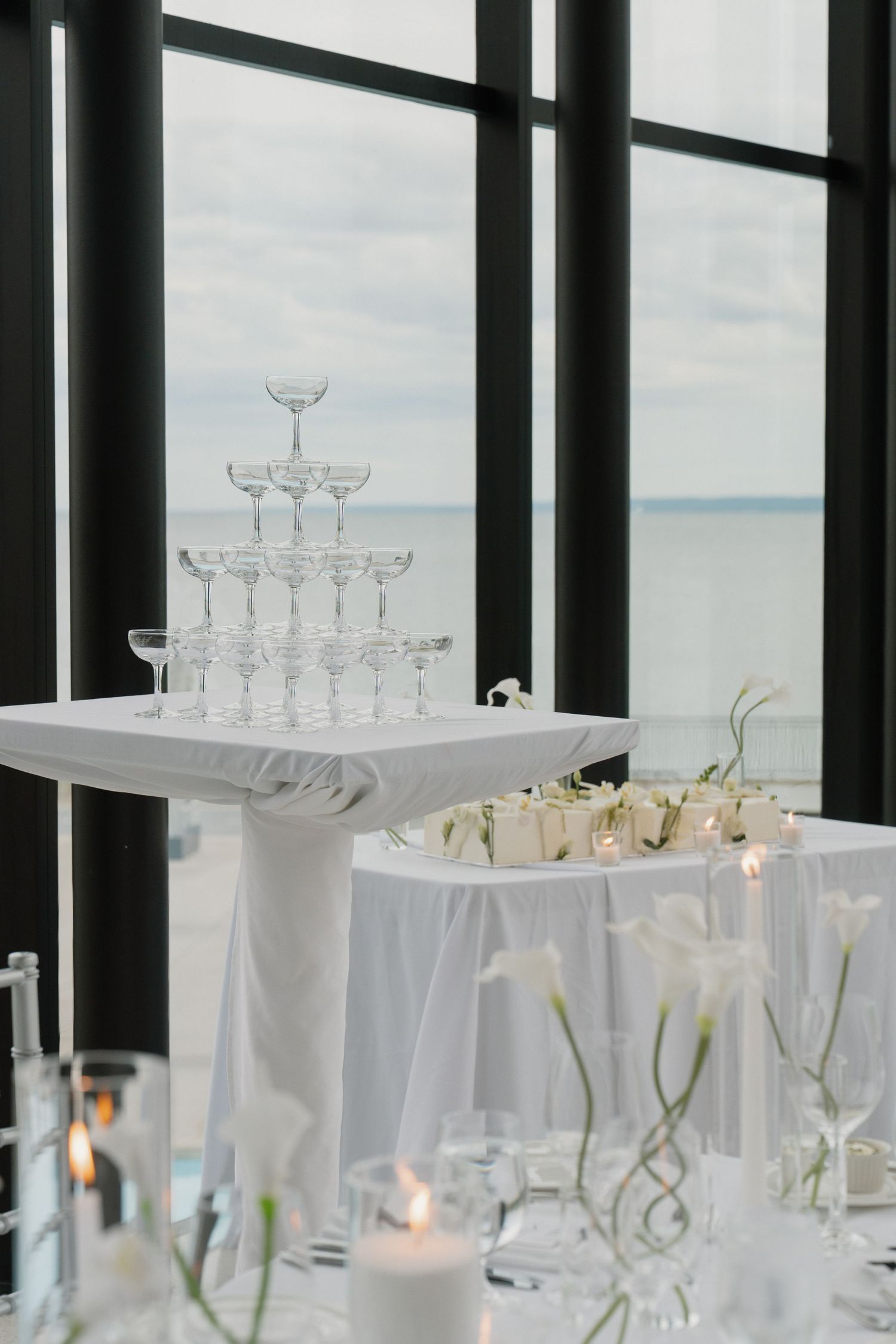 Elegant champagne tower with empty coupe glasses at a wedding reception, set against floor-to-ceiling windows overlooking the ocean.
