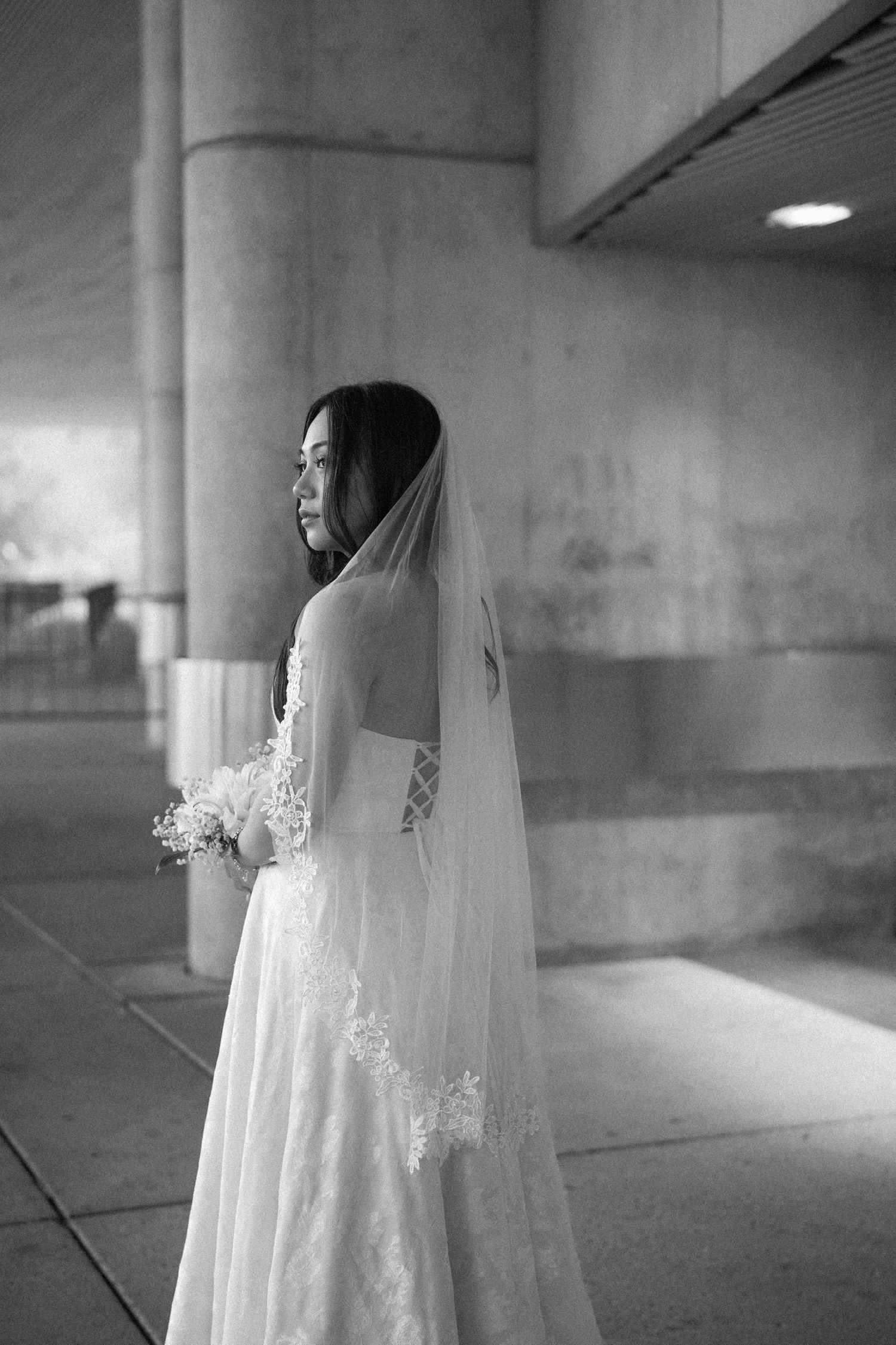 Black and white photo of a bride in a lace wedding dress and veil holding a bouquet near concrete pillars.