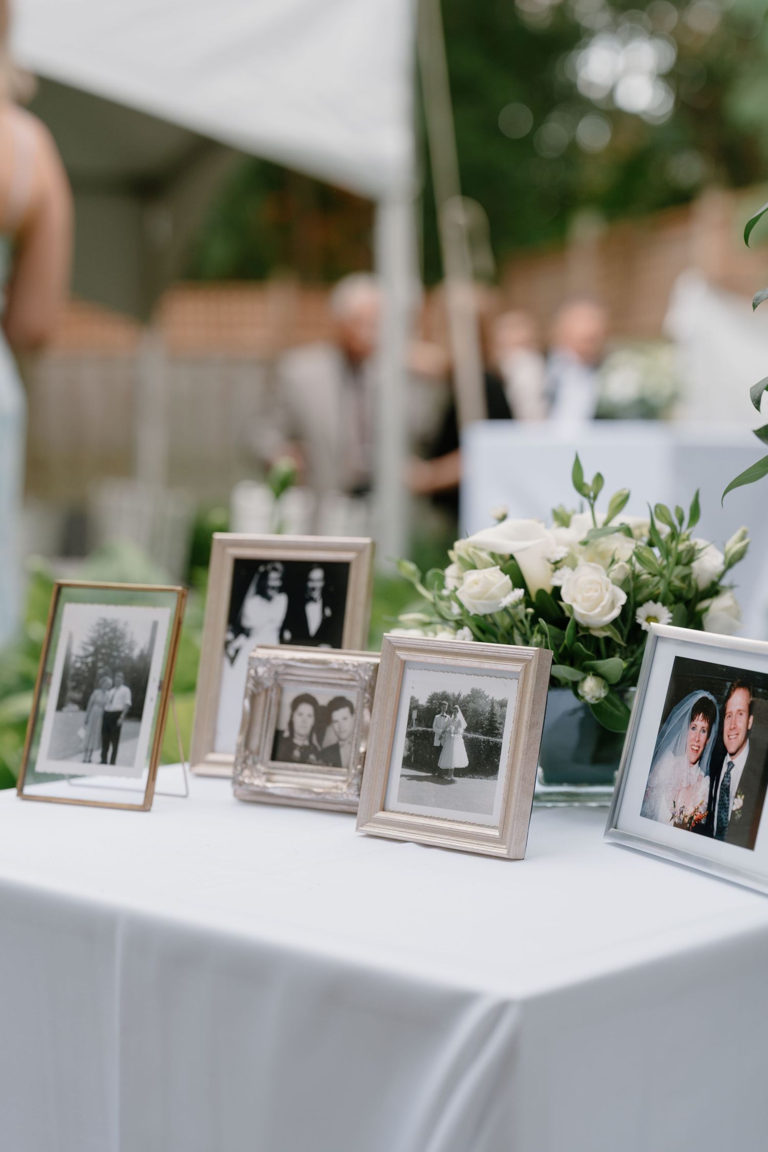 Framed black and white wedding photos displayed on a white tablecloth with white roses at an outdoor wedding reception.