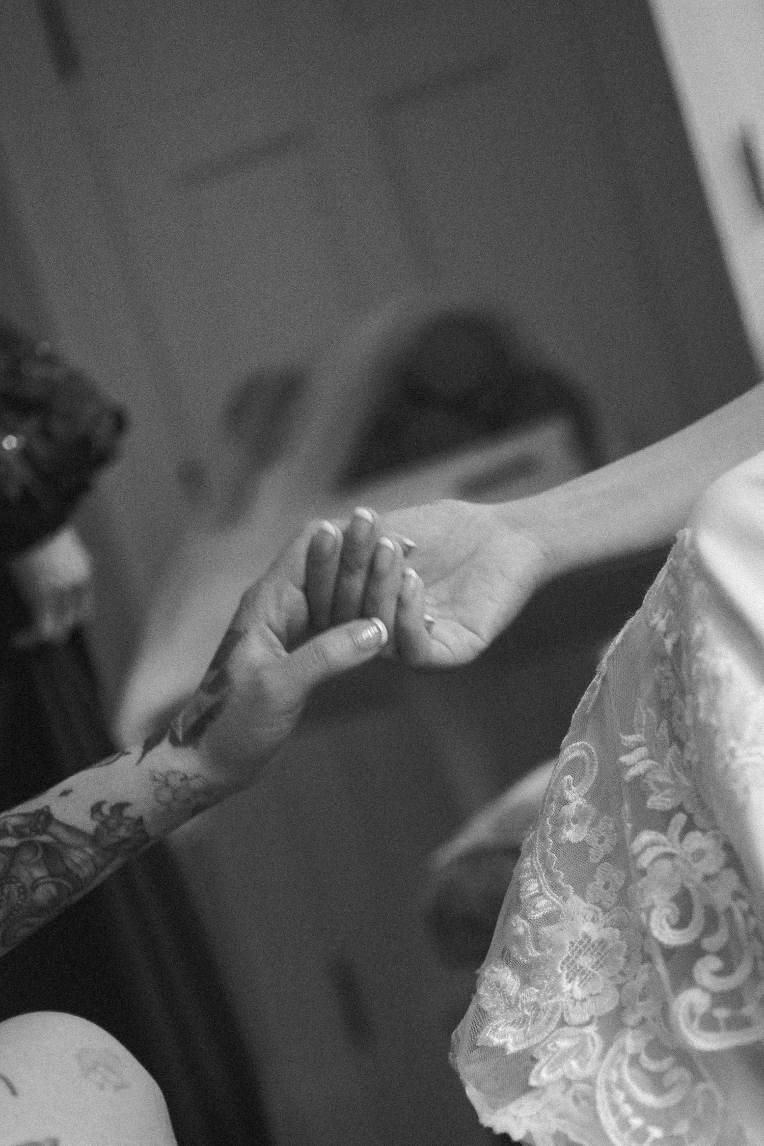 Black and white photo of a tattooed hand holding a bride's hand in a lace wedding dress.