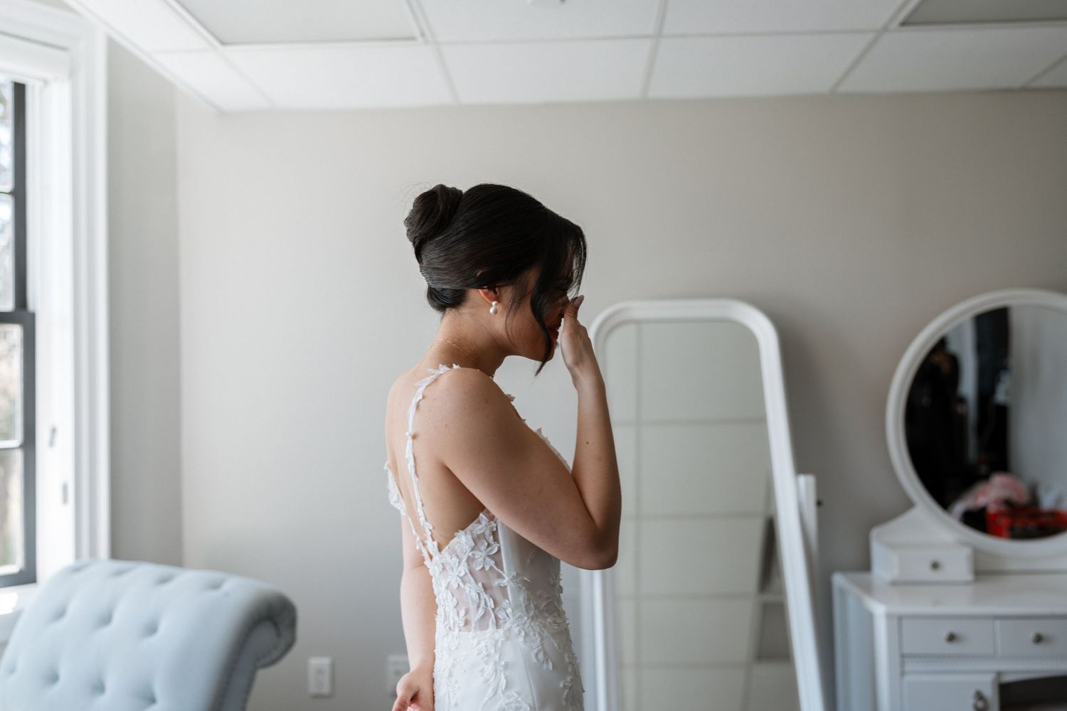 A bride wearing a delicate lace wedding dress stands in front of a mirror in a bright room with white furniture.