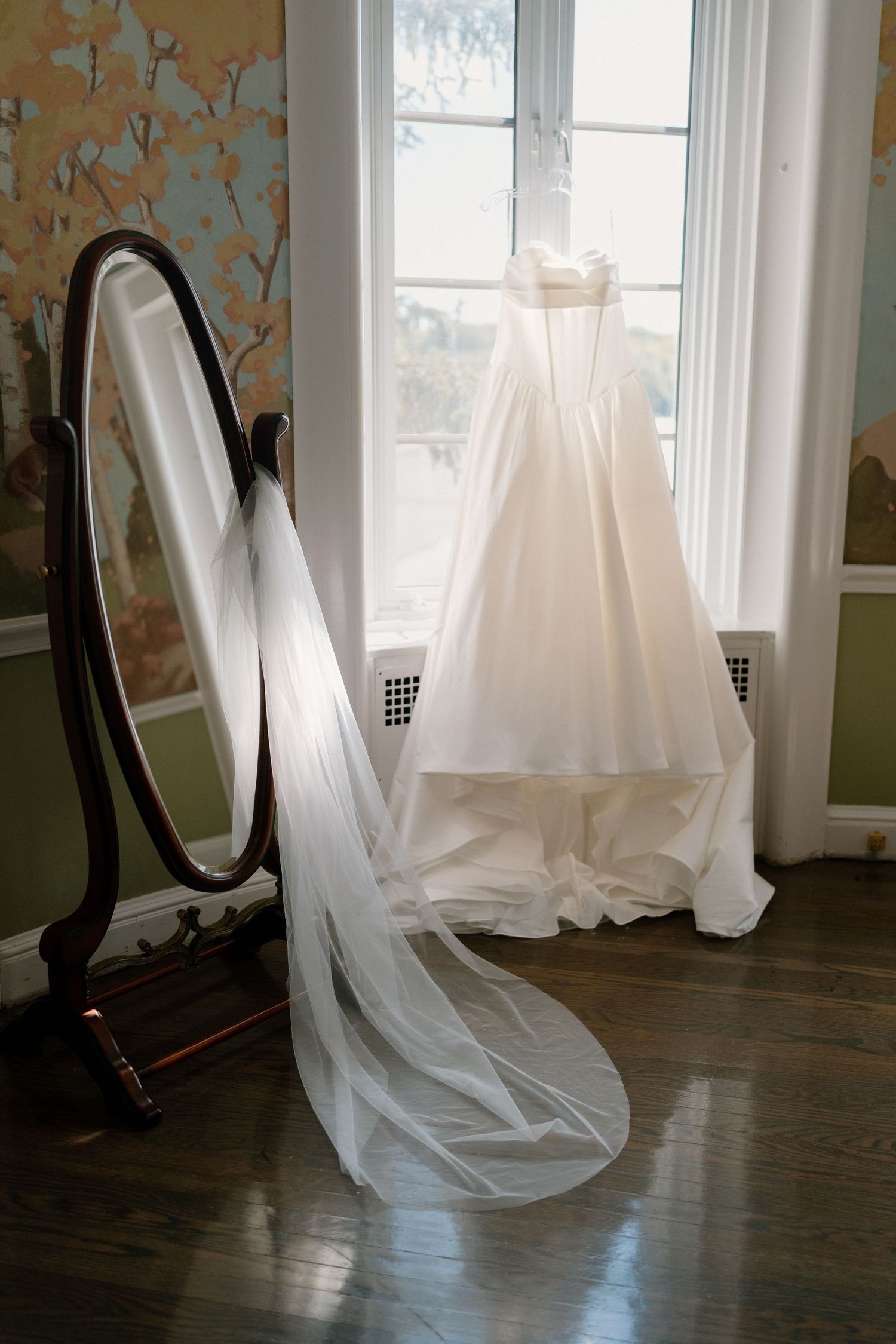 Elegant white wedding dress with long veil displayed near a window beside a vintage oval floor mirror in a decorated room.