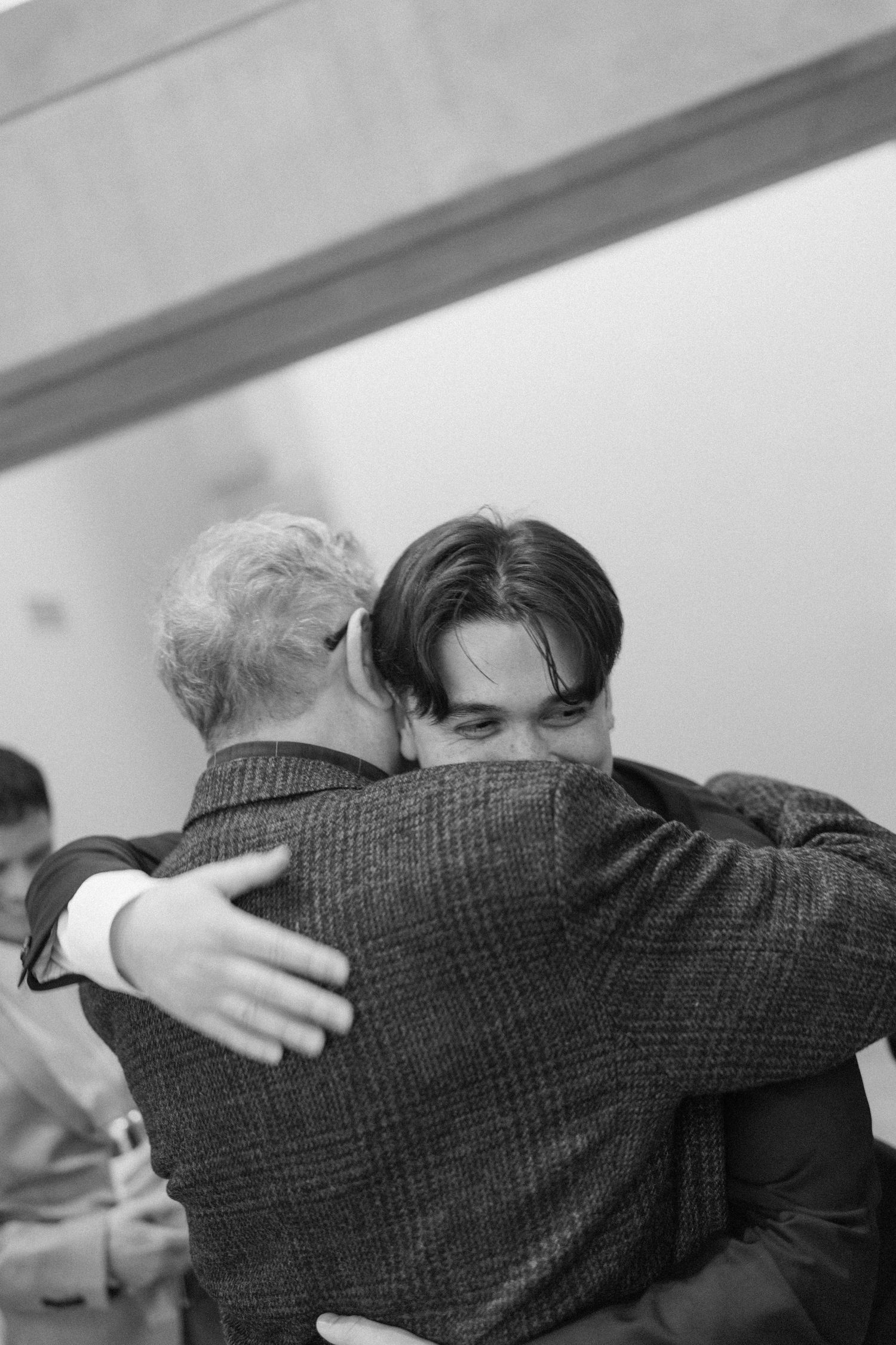 Black and white photo of two men sharing a heartfelt hug at a formal gathering, one with dark hair embracing an older gentleman.