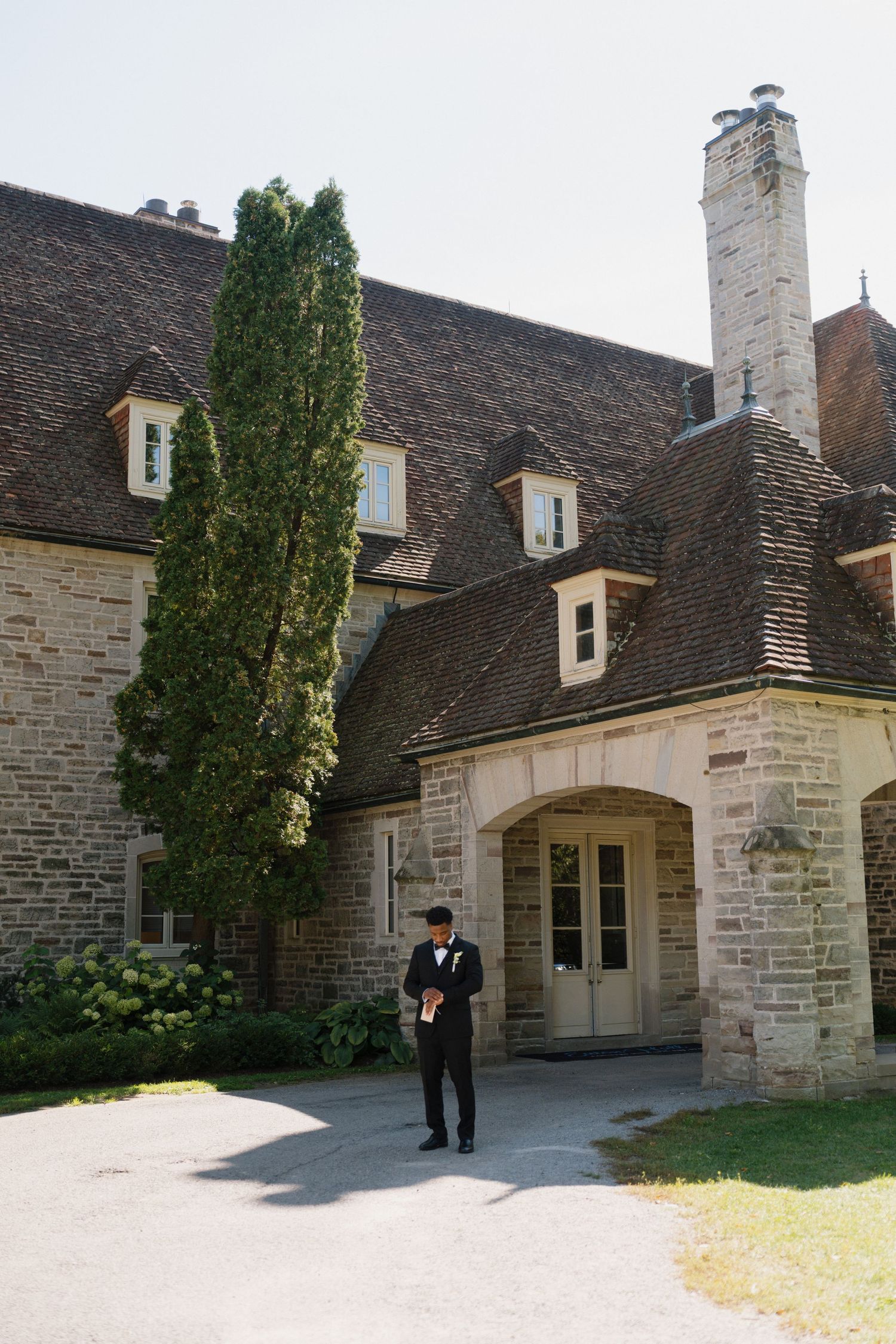 Groom in black tuxedo standing outside elegant stone mansion wedding venue with tall trees and arched doorway.